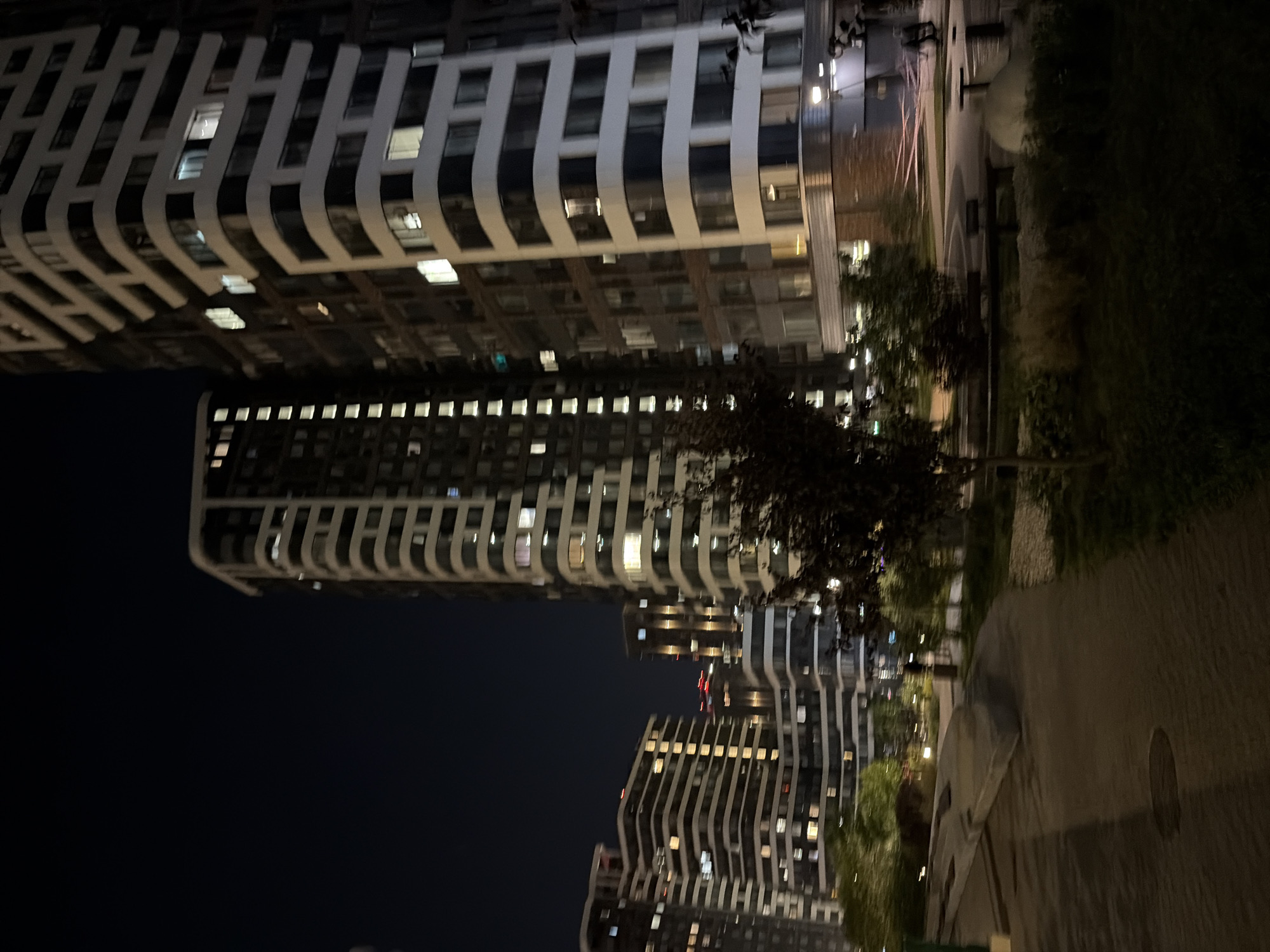 The image depicts a night view of a modern residential complex in Moscow, Russia. The scene is dominated by several tall buildings with illuminated windows, suggesting that they are occupied. The architecture features curved facades and a contrast between dark and light-colored materials. In the foreground, a paved pathway curves through a landscaped area with trees and grass. Streetlights are visible, casting a warm glow on the surroundings. The overall impression is one of a well-lit and contemporary urban environment at night.
