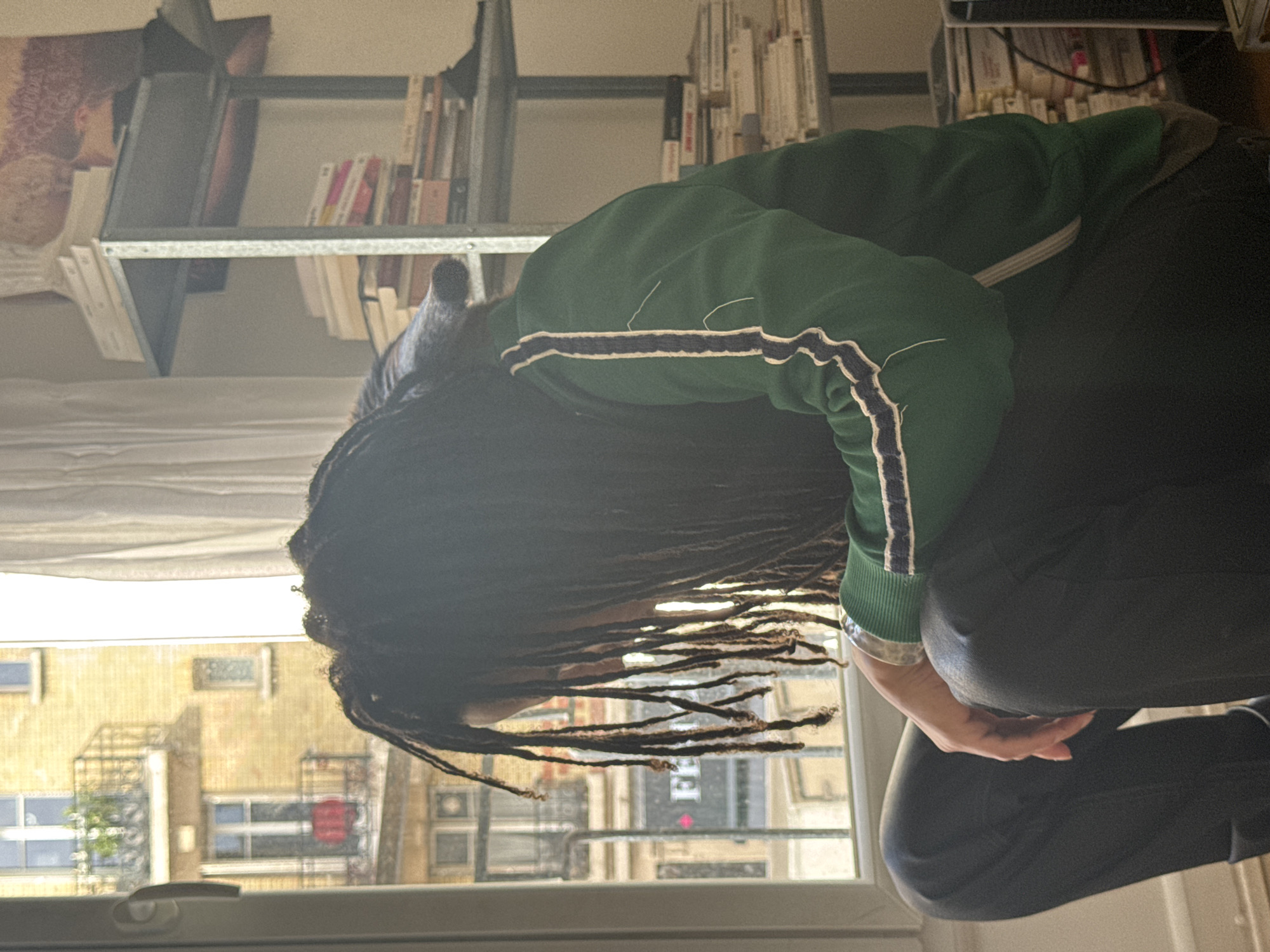 A person with long dreadlocks, wearing a green jacket with white stripes, is crouched by a window, with a view of buildings typical of Paris, France. The buildings are a beige color with many windows and balconies. A bookshelf with books is visible in the background. The person's face is obscured by their hair. They are wearing black pants and their hand is resting on their knee. The day appears overcast.