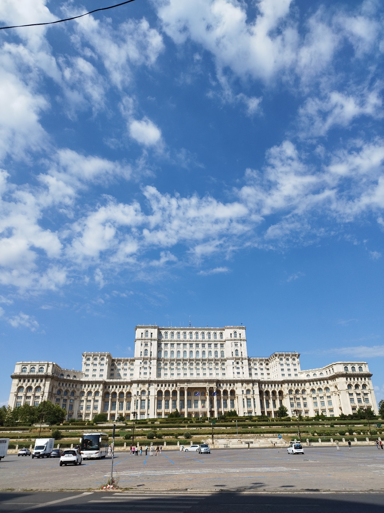 The image depicts the Palace of the Parliament in Bucharest, Romania, under a bright blue sky with scattered white clouds. The massive, neoclassical building dominates the background, showcasing its symmetrical facade, numerous windows, and grand architectural details. In the foreground, a paved area serves as a parking lot or plaza, with several vehicles including cars, vans, and a bus. A few people are visible walking in the plaza, and some are near the parked vehicles. The scene captures a sunny day in the city, with the imposing structure of the Palace of the Parliament as the central subject. The image is taken from a slightly low angle, emphasizing the scale of the building.