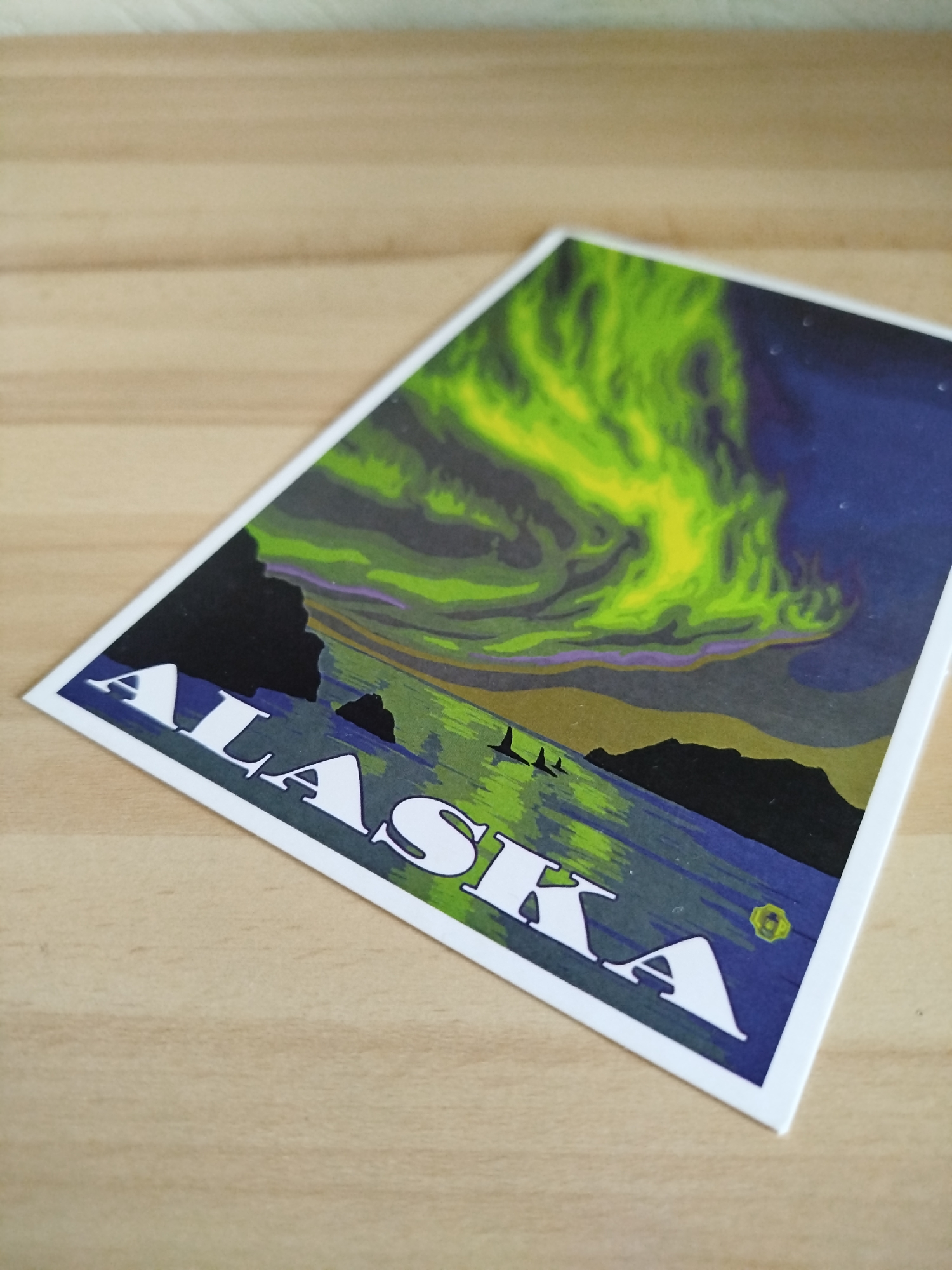 The image shows a rectangular postcard on a light-brown wooden surface. The postcard depicts a landscape scene with the word "ALASKA" printed in large, white, block letters along the bottom left side. Above the text is an illustration of a body of water reflecting the sky, with dark landmasses lining the horizon. In the sky, there's a bright green, swirling aurora borealis, contrasted by the deep blue of the night sky, dotted with a few stars. There is nothing to indicate that the image is related to the city of Varna, Bulgaria.