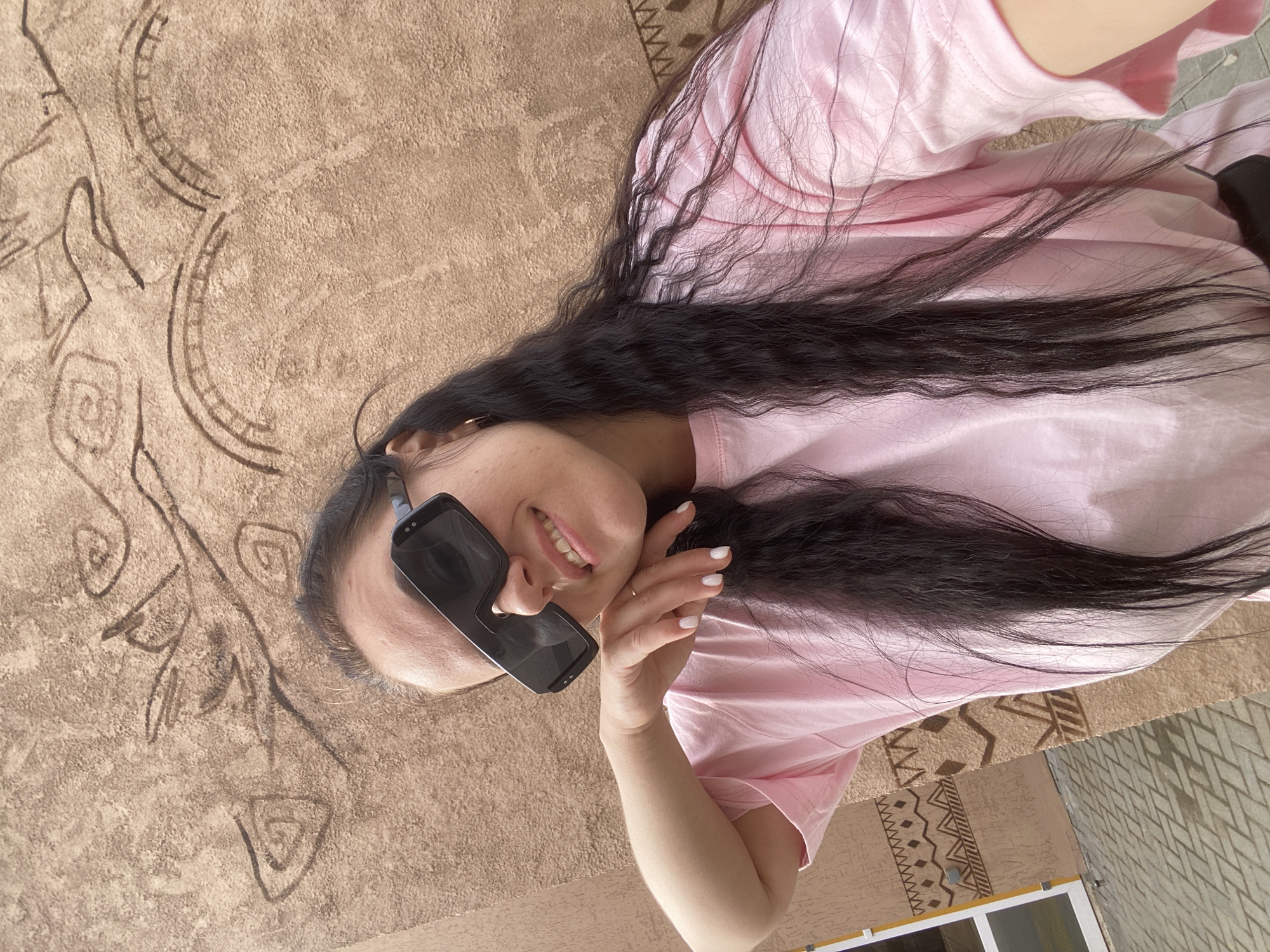 A young woman with long dark hair, wearing a pink t-shirt and black sunglasses, is posing for a photo. She is lying on a surface with an intricate, textured design that appears to be an exterior wall, possibly made of decorative concrete. The pattern has abstract, tribal-like drawings and geometric shapes. The background suggests an urban environment. The location is Perm, Russia.