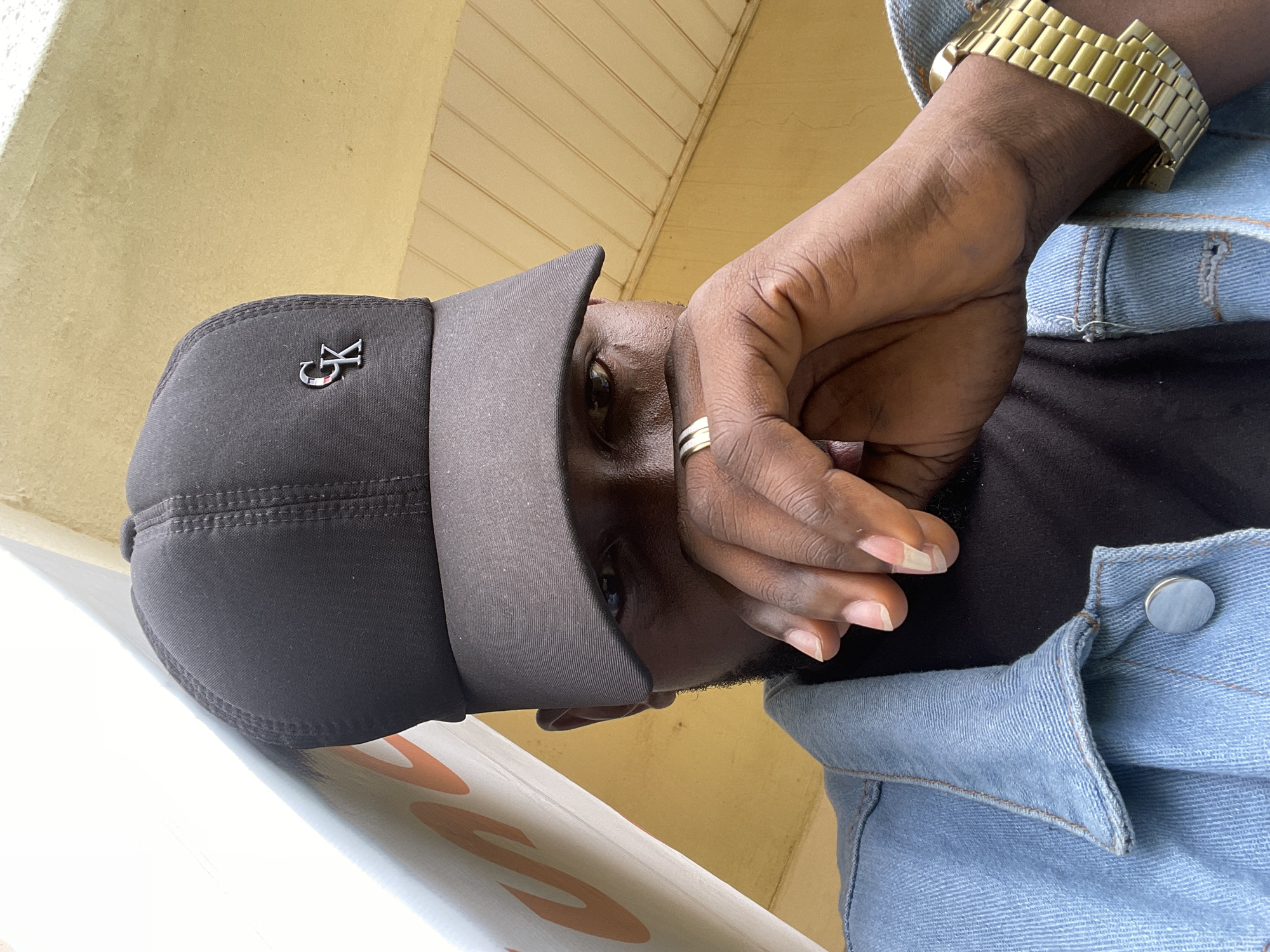 In Durumi, Nigeria, a person is seen from a low angle, wearing a black baseball cap with a silver "CK" logo. Their face is partially obscured by their hand, which is resting near their mouth. They are also wearing a denim jacket over a black t-shirt and a gold-colored watch on their left wrist. The background consists of a yellow wall and a white structure with orange lettering.