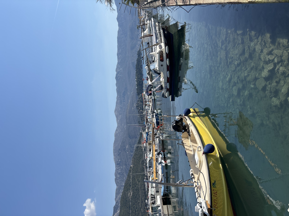 The scene depicts a clear, sunny day at a marina in Budva, Montenegro, with numerous boats moored in translucent water. In the foreground, positioned prominently on the left, is a bright yellow, open-hull motorboat with a black outboard engine, featuring "BD 310" and a fish graphic on its hull. Two dark blue cylindrical fenders are attached to its port side. To the right, in the midground, another motorboat with a white cabin and dark blue lower hull, labeled "BD 53," is also moored, displaying a smaller outboard motor and a stern railing. Both boats cast clear reflections onto the water, which is notably clear, revealing rocky formations on the seabed.
The marina extends into the background, populated by an array of various motorboats and sailboats, all appearing to be docked. A concrete or stone quay edge is visible along the right side of the image. The immediate background features the dark, treed slopes of coastal hills, which ascend to a range of rugged, grey-blue mountains under a predominantly clear, light blue sky. A single small white cloud is present in the far upper left. No people are visible, and no discernible actions or interactions are occurring.