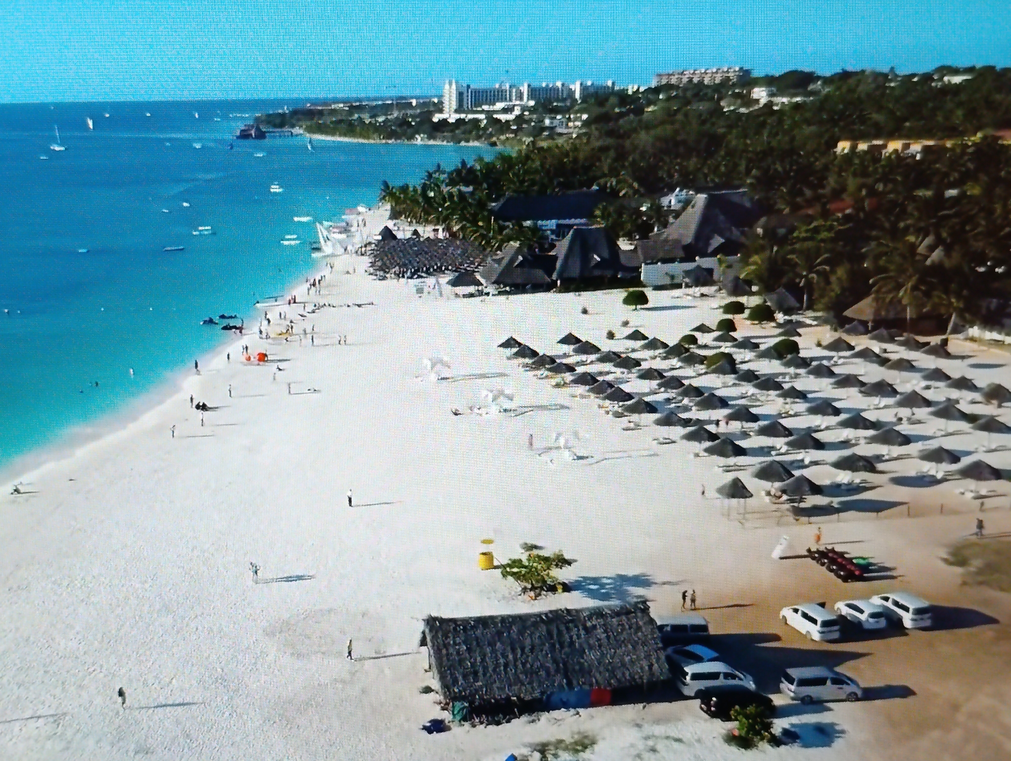 This aerial image depicts a crowded tropical beach with white sand and clear turquoise water. Numerous thatched-roof umbrellas are set up on the beach, providing shade for lounge chairs. Several people are visible on the beach, some walking, some sitting, and others wading in the water. In the background, a resort with multiple buildings and palm trees is situated along the coastline, with a cityscape visible further inland. Several boats are anchored in the ocean. The scene suggests a popular tourist destination with recreational activities taking place. The location is identified as Dubăsari town council, Moldova.
