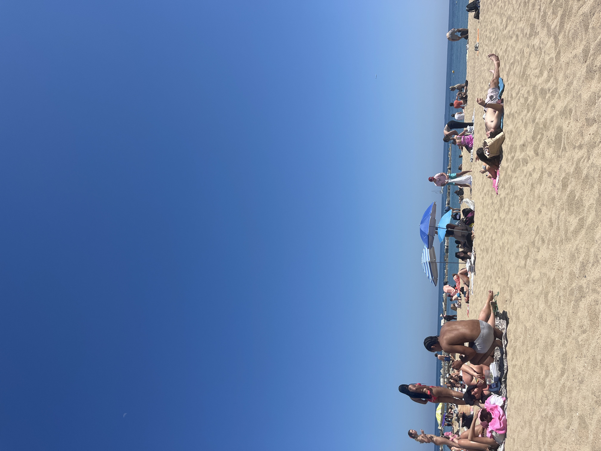 The image depicts a sunny day at a crowded beach in Barcelona, Spain. A large expanse of clear blue sky dominates the frame. The beach is sandy and filled with people sunbathing, sitting, and walking. There are several colorful beach umbrellas scattered throughout the scene. The sea is visible as a thin strip in the background.