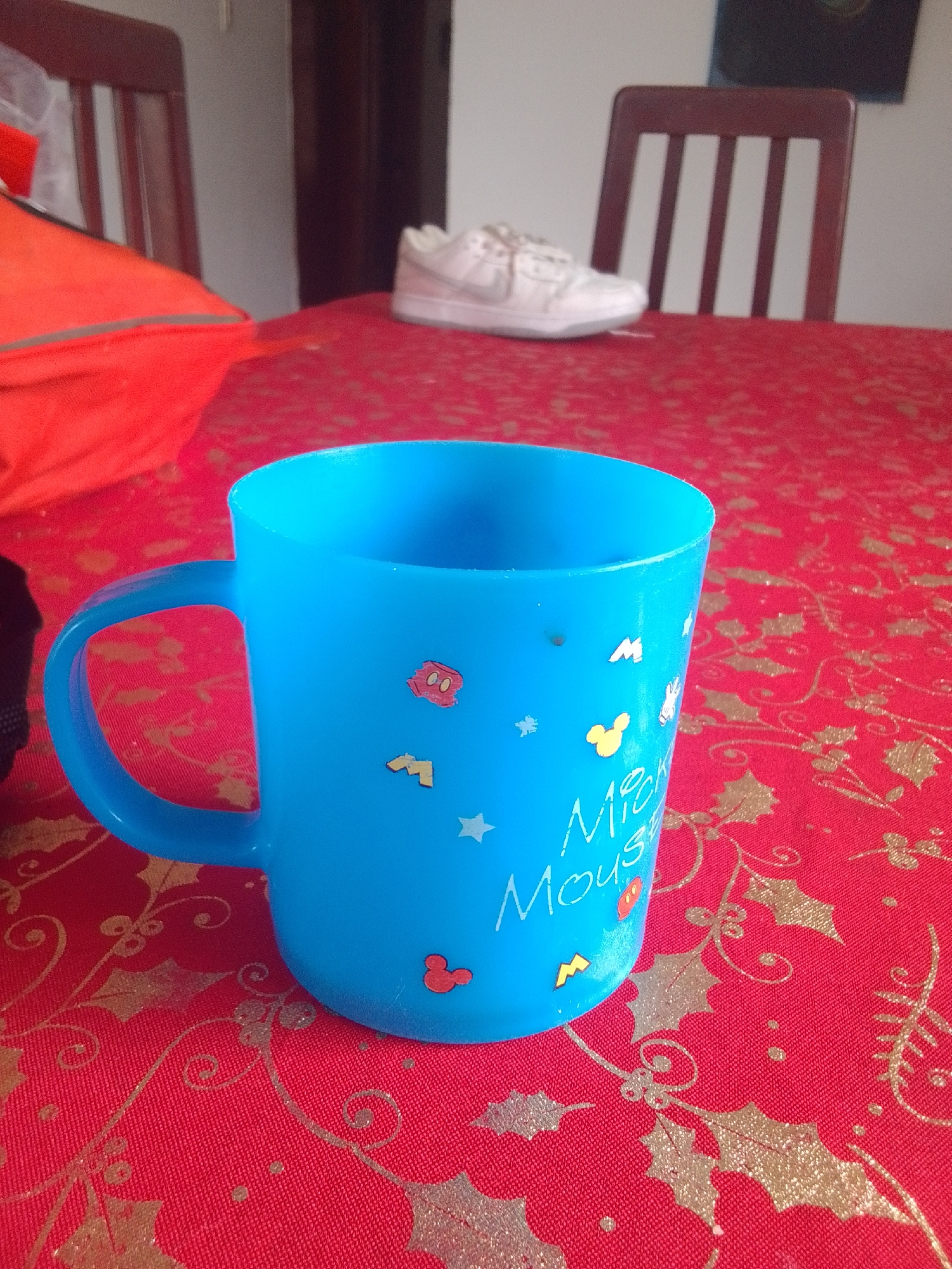 A blue plastic mug with Mickey Mouse decals sits on a red tablecloth with a gold holly pattern. The mug has a handle and is decorated with various cartoon characters and stars. In the background, two wooden chairs with white slats are visible. A pair of white sneakers is placed on a surface behind the mug. The scene appears to be indoors, potentially in a home setting.