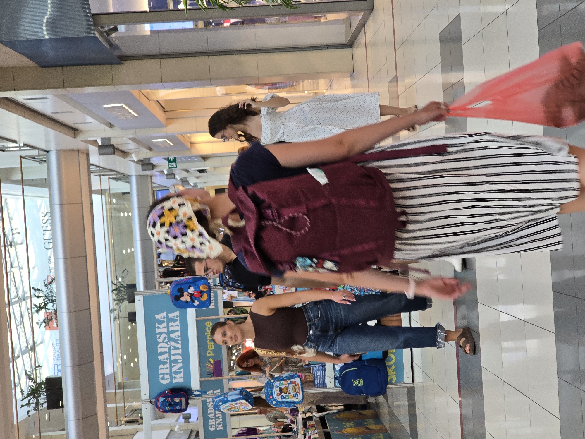 In Podgorica, Montenegro, within a modern shopping mall, a woman with a patterned headscarf and a burgundy backpack walks away from the camera, carrying a red plastic bag. She is wearing a striped skirt and a dark top. To her left, a woman in a dark brown sleeveless top and blue jeans walks towards the left of the frame, carrying a light-colored purse. In the background, a woman in a white polka dot dress is walking towards the right. A store sign with "GRADSKA KNJIŽARA" (City Bookstore) is visible on the left, displaying several children's backpacks. The mall's interior features a tiled floor and architectural elements characteristic of a commercial space.