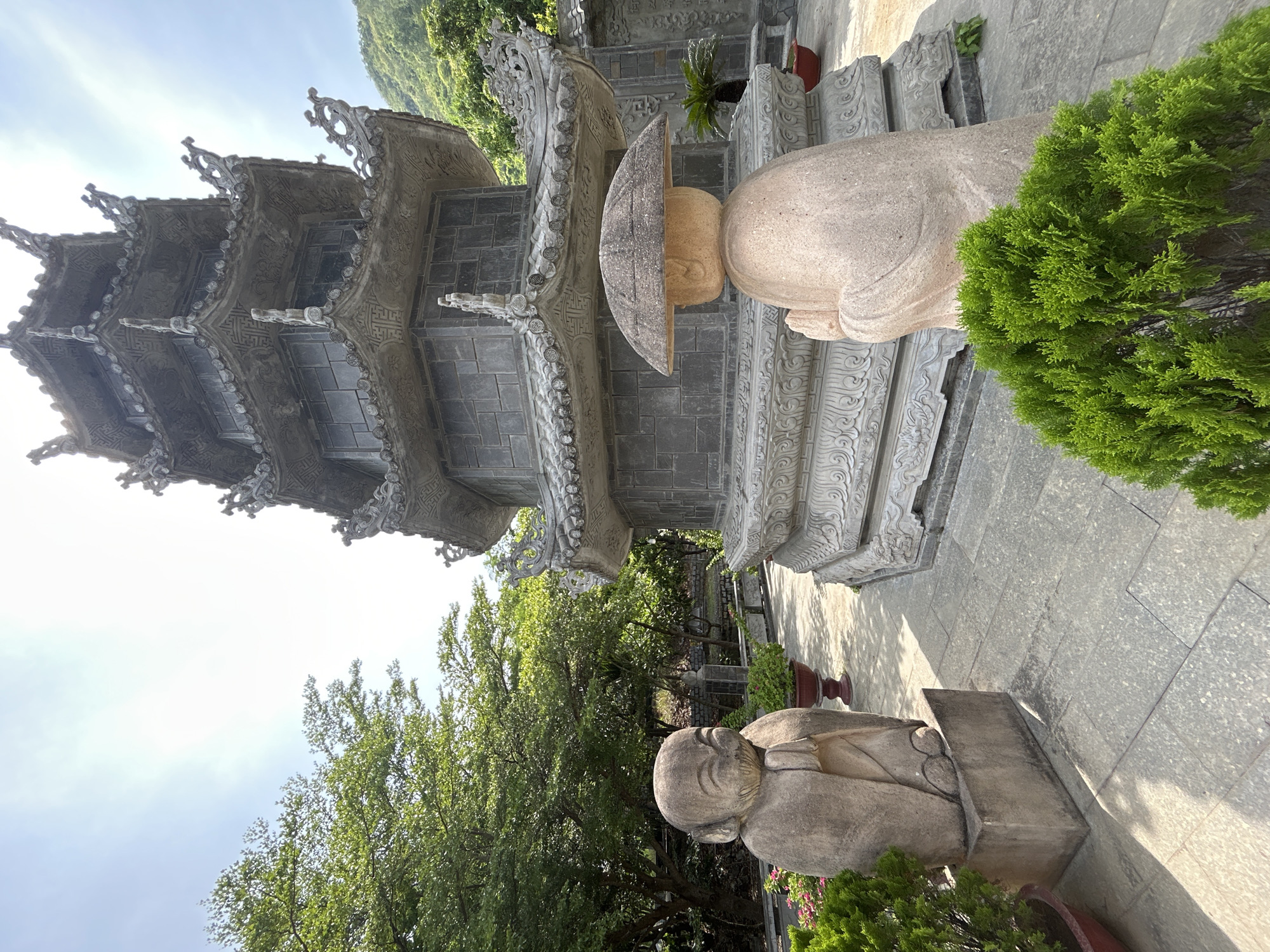 This is a full, eye-level, outdoor shot of a stone pagoda and two stone statues in the city of Nam Nha Trang Ward, Vietnam. In the foreground, two stone statues stand on either side of a paved walkway. The statue on the left is shorter, with a round face and hands clasped in front of it. It is smiling with its mouth open. The statue on the right is taller, and its face is obscured by a wide, conical hat. Its hands are also clasped in front of it. Both statues appear to be made of sandstone. Behind the statues, a large, multi-tiered pagoda dominates the center of the frame. The pagoda is made of dark gray stone, with intricate carvings and decorative elements on each tier. The pagoda's roof is tiled and curves upwards at the edges. The background consists of lush green trees and a hazy, bright sky with some clouds. The ground is paved with large, rectangular stone tiles, and there are some potted plants scattered around the area. Sunlight illuminates the scene, casting shadows on the ground. The overall atmosphere is one of tranquility and spirituality.