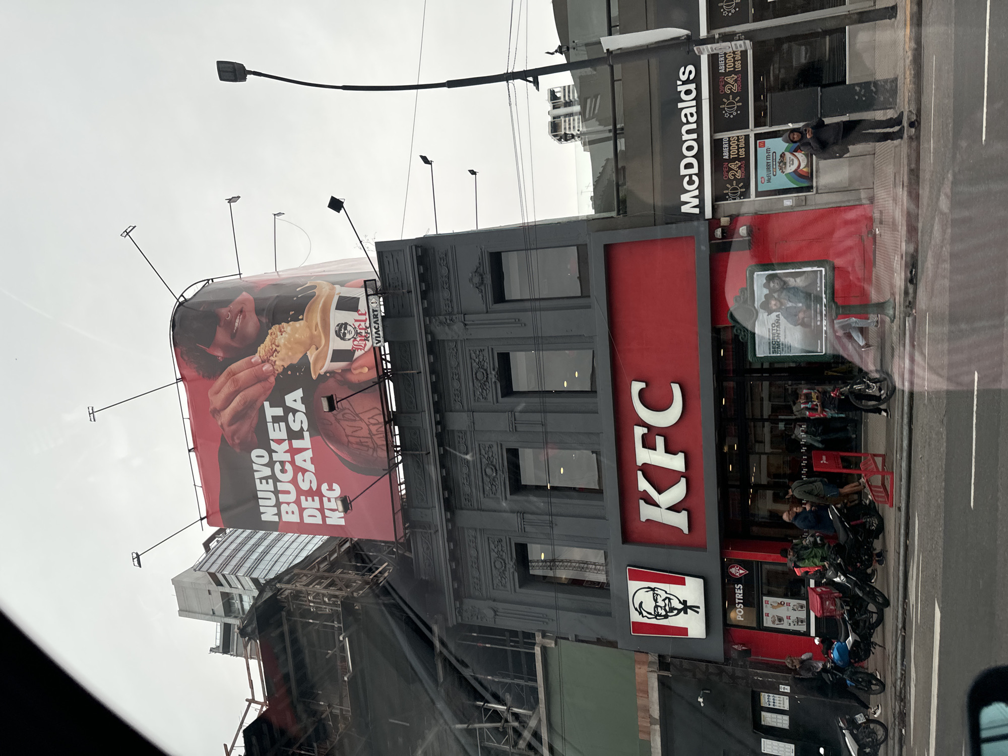 The image captures a street scene in Buenos Aires, Argentina, featuring fast-food restaurants and urban infrastructure. A KFC restaurant dominates the lower right, with a large "KFC" sign and a smaller sign displaying Colonel Sanders' face. Above the KFC, a large billboard advertises a "Nuevo Bucket de Salsa KFC." To the right of the KFC is a McDonald's restaurant, recognizable by its signage and branding. People are visible near the entrances of both restaurants. Several motorcycles are parked in front of the KFC. Above the restaurants, utility poles and wires are seen against an overcast sky. Additionally, scaffolding and construction materials are visible on the left. A building under construction is partly obscured by the billboard.