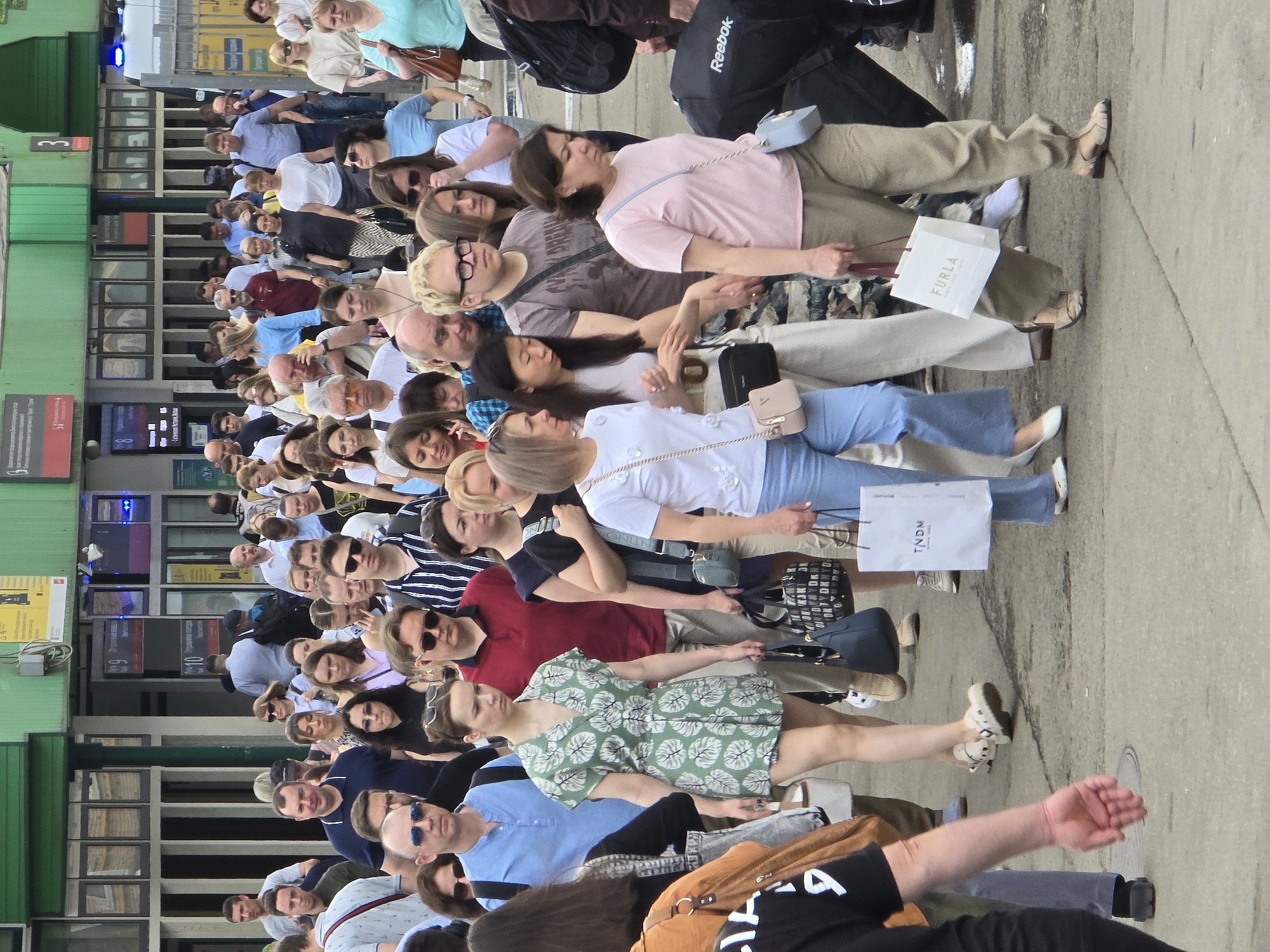 A large queue of people stands outside what appears to be a train station in Moscow, Russia. The station has green paneling on the exterior with visible signs in Russian. The people are diverse in age and attire, mostly dressed in casual summer clothing. Some individuals are carrying shopping bags from brands such as Furla and TNDN. There is a Reebok bag visible. People are standing closely together, and the crowd extends significantly. The setting is an urban environment with a paved sidewalk or street.