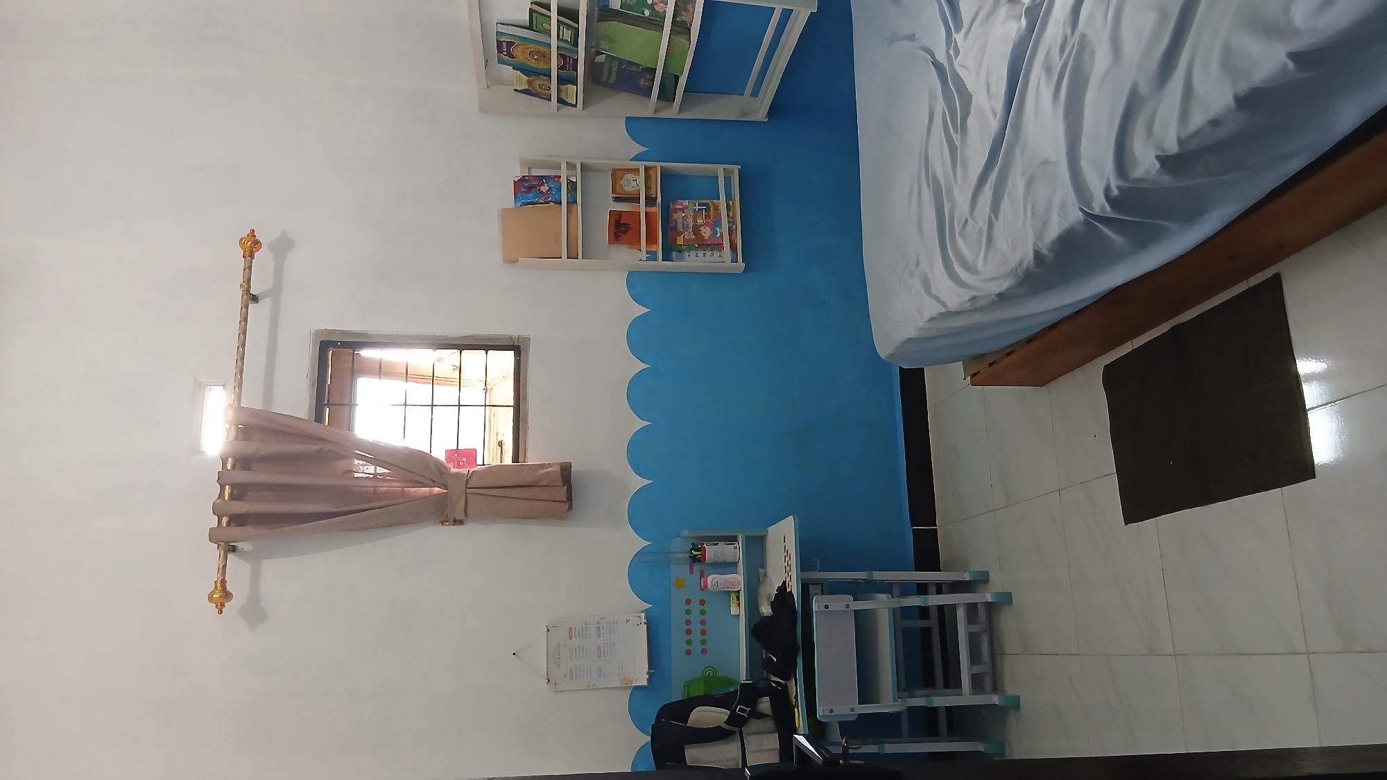 The image depicts a bedroom interior with a bed, a desk, and bookshelves. The room is painted white and blue with a scalloped border along the lower half of the walls. A window with brown curtains is visible on the upper portion of the left wall. A small dark rug lies on the tiled floor next to the bed. The desk on the left has a backpack and some stationery items on it. Two white wall-mounted bookshelves are on the right wall, filled with books. The overall impression is of a tidy, organized room, likely a child's bedroom, possibly located in Klambir Lima Kebun, Indonesia, given the context.