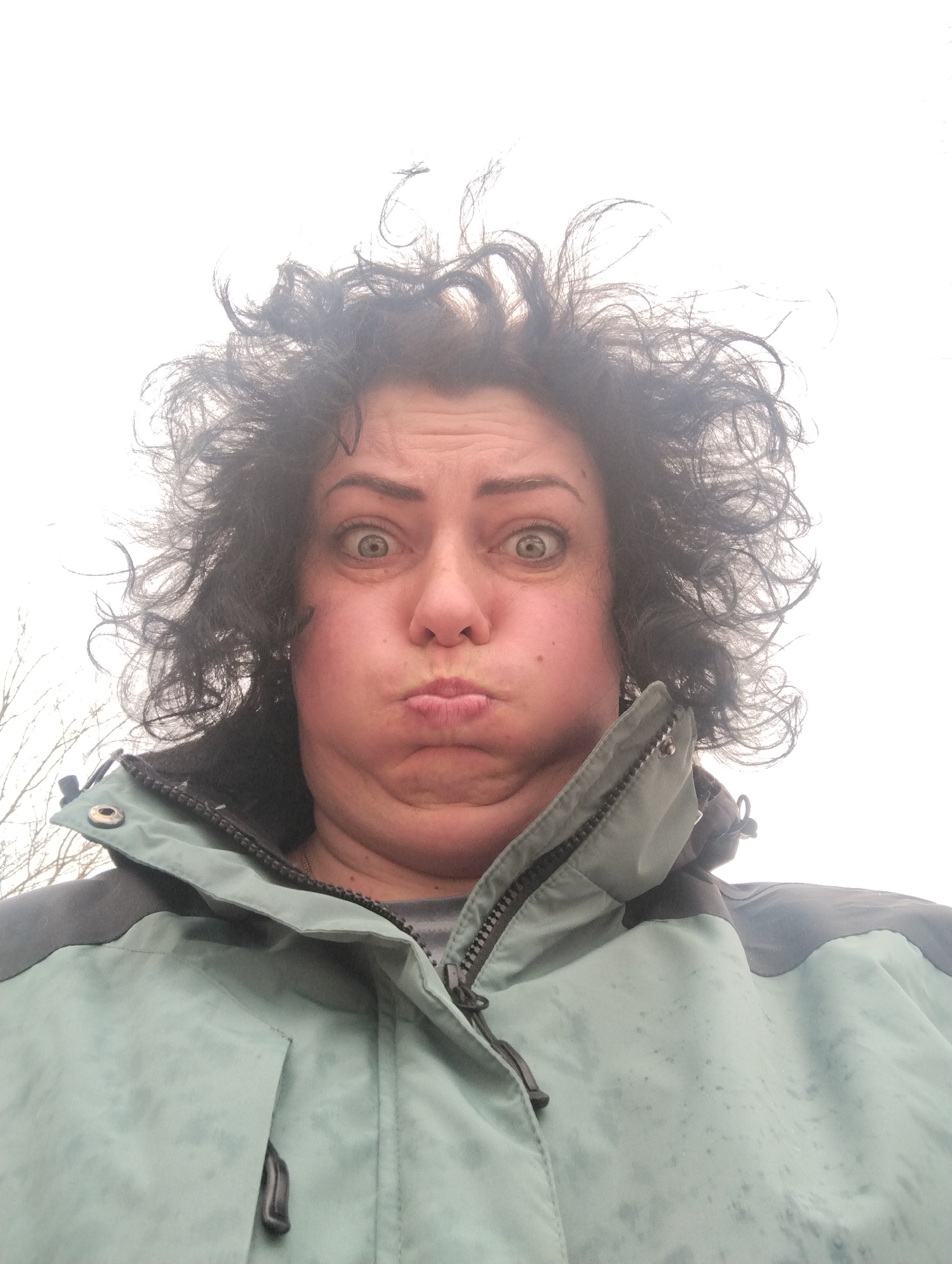 A woman with dark, curly hair and light skin is making a pouting expression directly into the camera. Her eyes are wide. She is wearing a light blue and dark gray jacket. The background is white and indistinct, with bare branches visible in the lower left corner. The location is Grishanki, Russia.