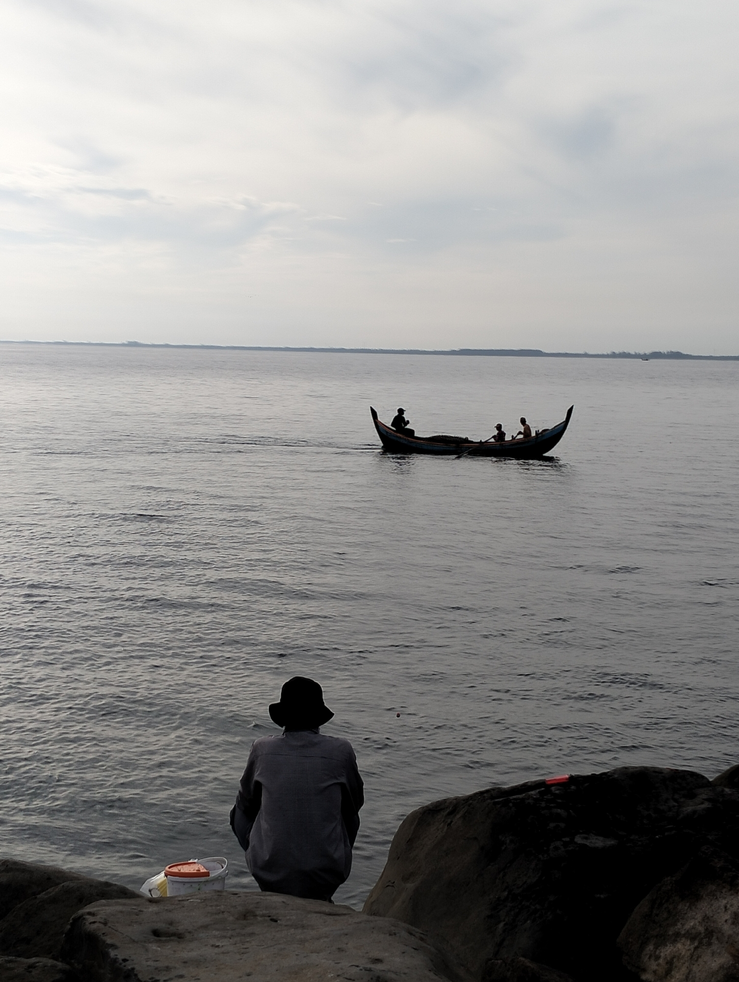 A calm ocean view, likely Lhokseumawe, Indonesia. A person wearing a hat and a checkered shirt sits on dark rocks at the edge of the water, facing away from the viewer. A white bucket with an orange lid sits next to the person. In the distance, a long, narrow boat with three occupants moves across the water. The sky is overcast. A thin strip of land is visible on the horizon.