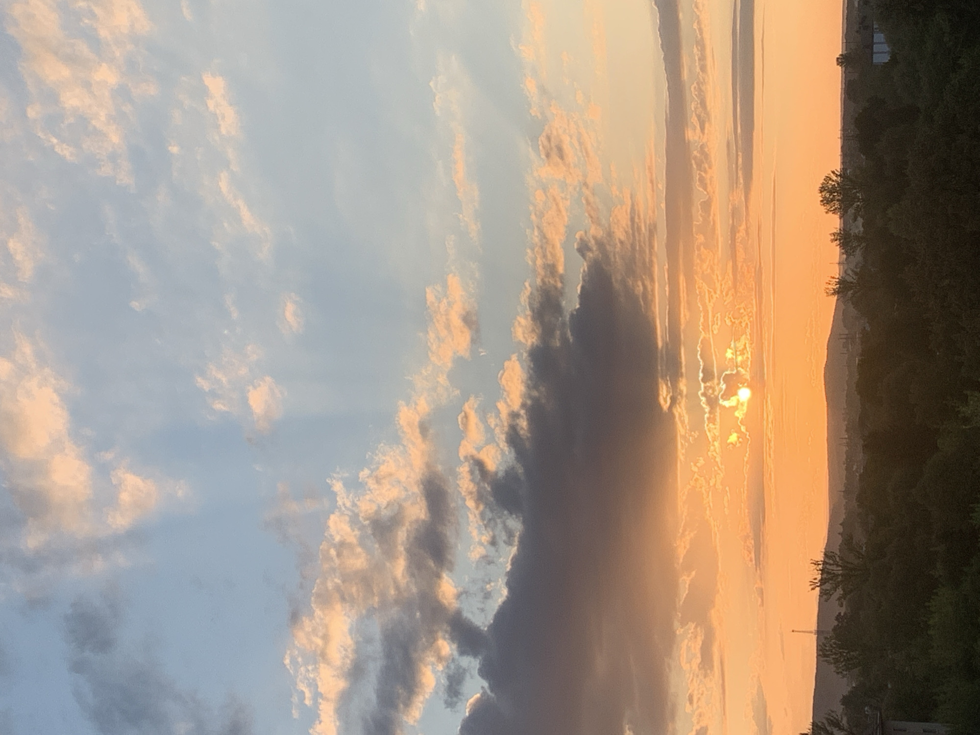 This is a photograph of a sunset over Kolomyia, Ukraine. The sky is filled with clouds tinged with orange and pink from the setting sun. The sun itself is a bright circle peeking through a gap in the clouds, casting rays of light downwards. Below the sky, a dark silhouette of trees and hills stretches across the horizon, with some buildings and a radio tower visible in the distance. The overall impression is one of a serene, natural landscape at the end of the day.