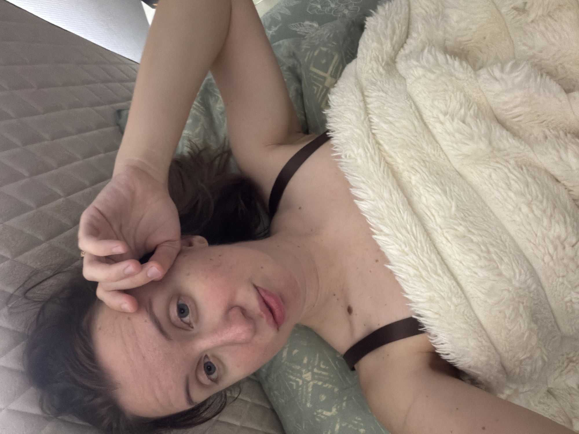 A young woman with brown hair and blue eyes is lying in bed, looking at the camera. She is wearing a dark bra and is covered by a fluffy white blanket. Her right hand is resting on her forehead. The background appears to be a bedroom with a patterned pillow and a textured headboard. The lighting suggests it is indoors, possibly in the morning or evening. The specific location in Bila Tserkva, Ukraine, is not discernible from the image.