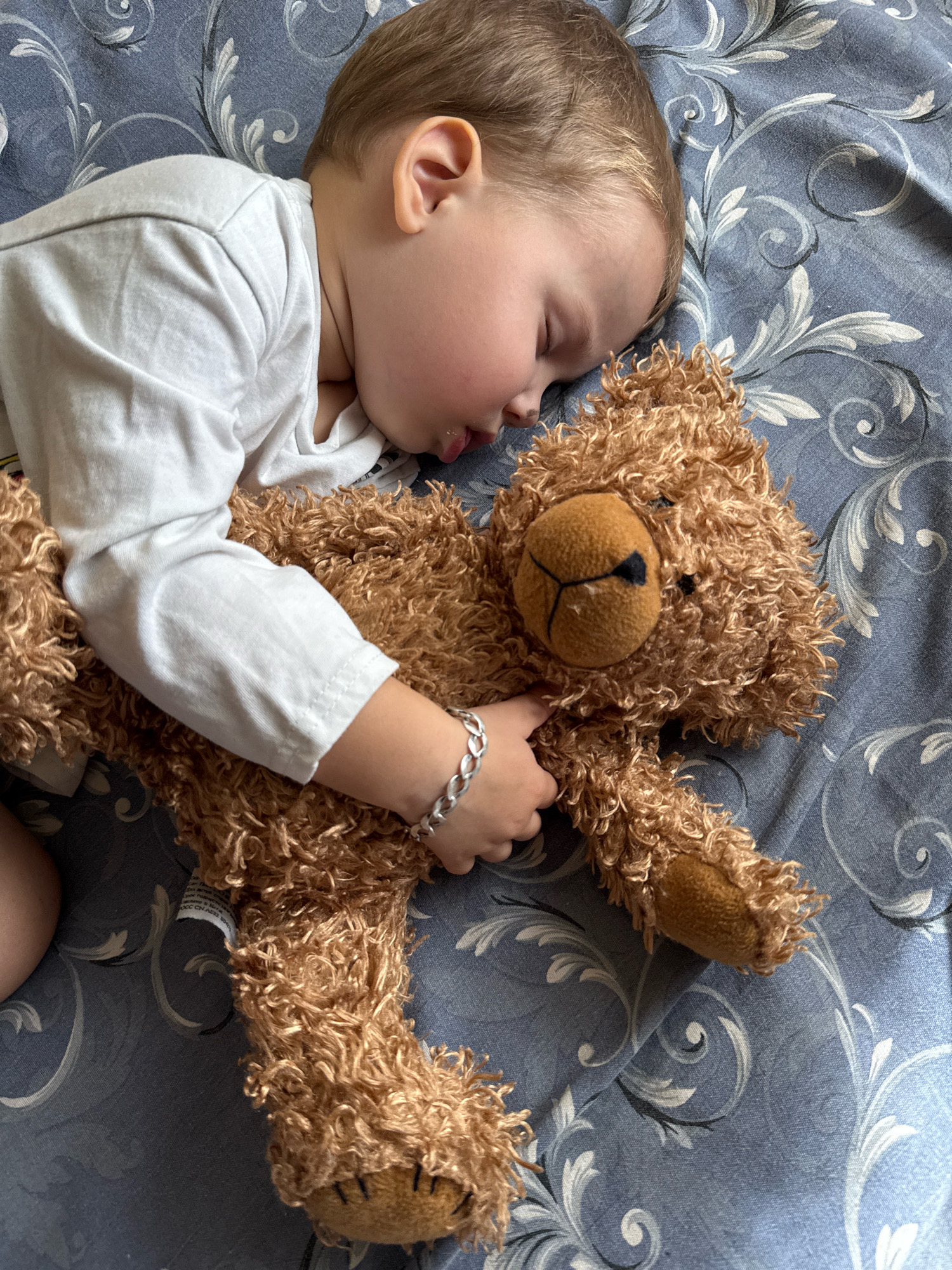 A toddler, estimated to be between 2 and 3 years old, sleeps soundly on a blue, patterned bedspread. The toddler is wearing a white long-sleeved shirt. An arm is wrapped around a brown, fuzzy teddy bear, which is also lying on the bedspread. The teddy bear has a tan-colored snout with black stitching for its nose and mouth. A silver bracelet is visible on the toddler's left wrist. The scene is indoors, with the bedspread filling the frame. The location is not specified as being in Rusanovo, Russia, or any other city.
