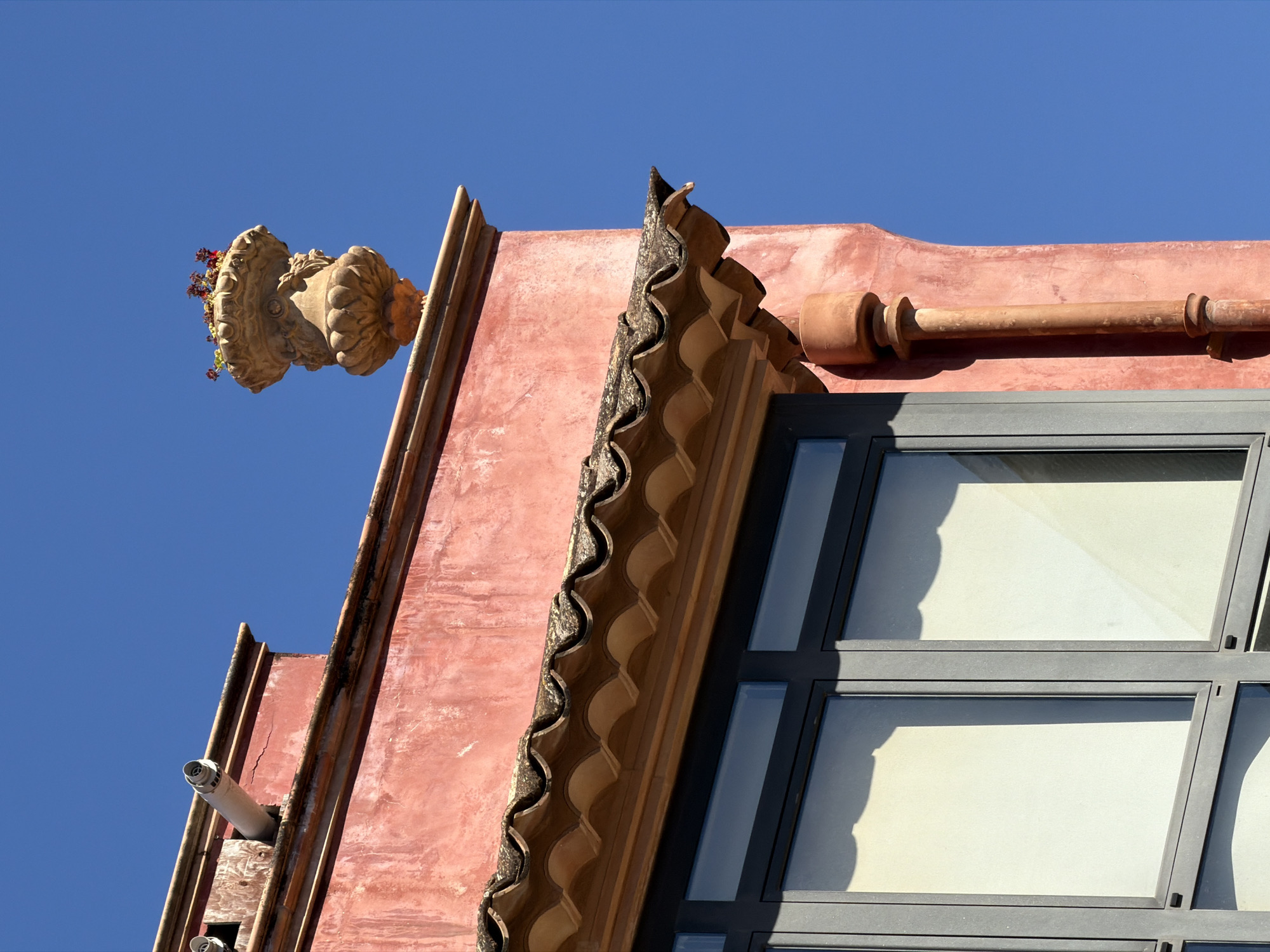The image shows an exterior view of a building in Manacor, Spain, against a clear blue sky. The building features a weathered salmon-pink stucco facade. A section of a tiled roof with curved, overlapping tiles is visible, extending over a window with a dark grey frame. The window has multiple panes, and reflections of light and shadows are present on the glass. A terracotta downspout runs vertically along the side of the building. At the top of the building, a decorative urn filled with small, red flowers sits on an ornate ledge. A white pipe, possibly a drainpipe or vent, protrudes from the building near the urn. The scene is characterized by architectural elements and a bright, sunny atmosphere.