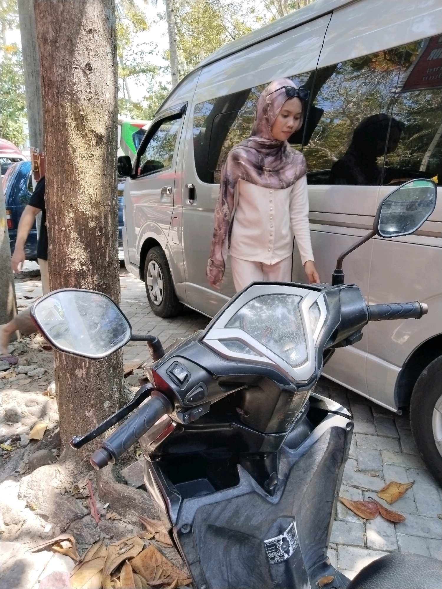 In Prigi, Indonesia, a person wearing a hijab and a light-colored top and pants stands next to a silver van, with sunglasses perched on their head. The person's hand is resting on the van's side. In the foreground, a black motorcycle is parked, its side mirror visible. The scene is outdoors, with a large tree trunk to the left of the frame. The ground is covered with fallen leaves and some gravel. The overall environment appears to be a roadside or parking area.