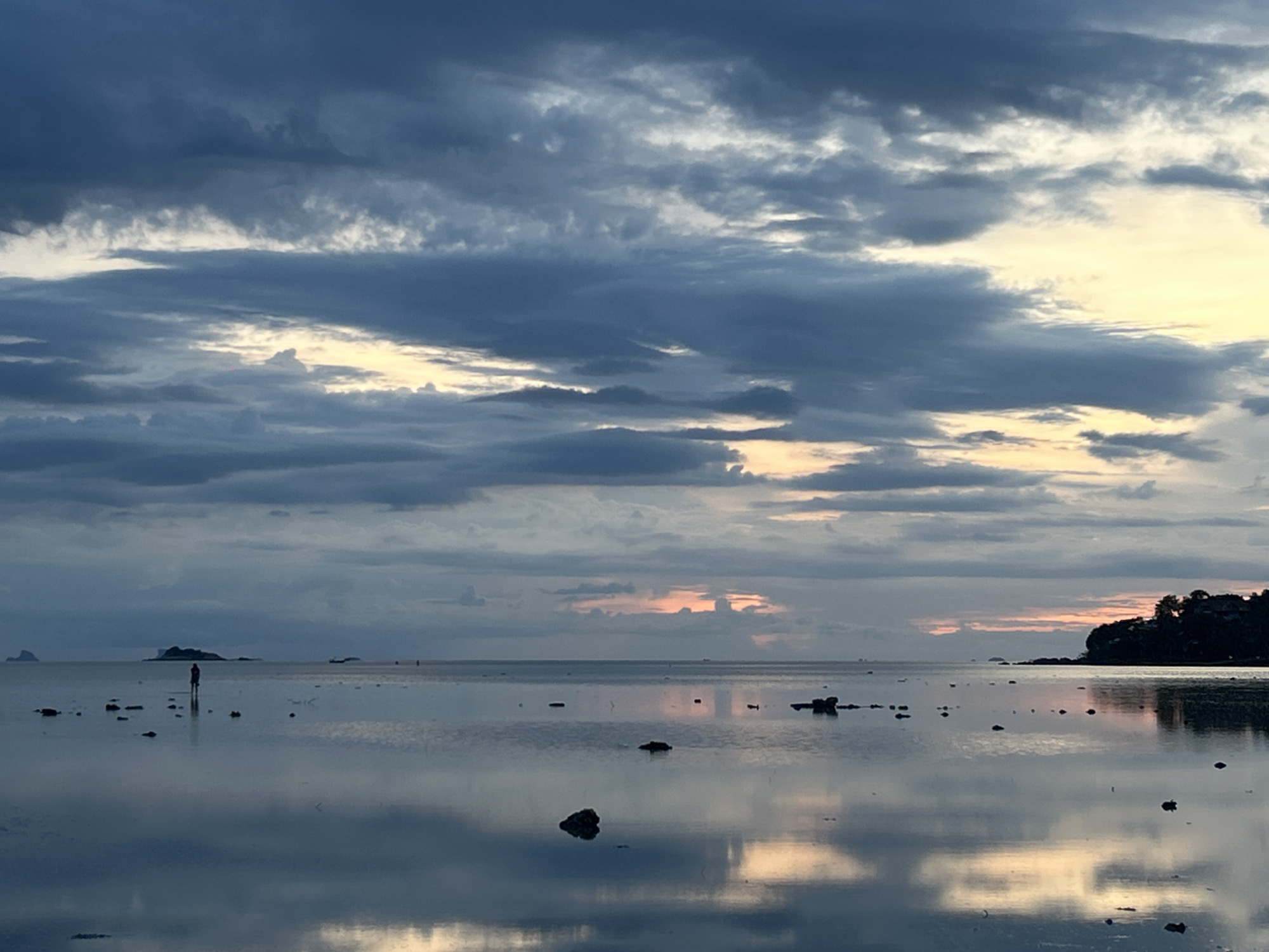 The image depicts a serene seascape at dusk or dawn on the island of Koh Phangan, Thailand. The sky is a canvas of dramatic, layered clouds, predominantly in shades of deep blue and grey, with streaks of pale yellow and soft pink near the horizon, suggesting the fading or emerging light of the sun. The sea is remarkably calm and reflective, mirroring the sky and creating a glassy surface broken only by the occasional dark rock or small island visible in the distance.
In the foreground and mid-ground, scattered rocks emerge from the shallow water, their dark forms contrasting with the muted tones of the sea and sky. The most prominent human subject is a solitary figure standing in the water, silhouetted against the reflective surface. This individual is positioned to the left of the frame, facing away from the viewer, and their presence adds a sense of scale and contemplation to the vastness of the scene.
To the right of the frame, a dark, silhouetted landmass, likely a coastline or another island, rises from the water. It is covered in dense vegetation and features structures that are indistinct in the low light. The reflection of this landmass is visible in the water below, further blurring the line between sea and sky. The overall impression is one of tranquility, natural beauty, and the quiet solitude often found in coastal landscapes at transitional times of day.