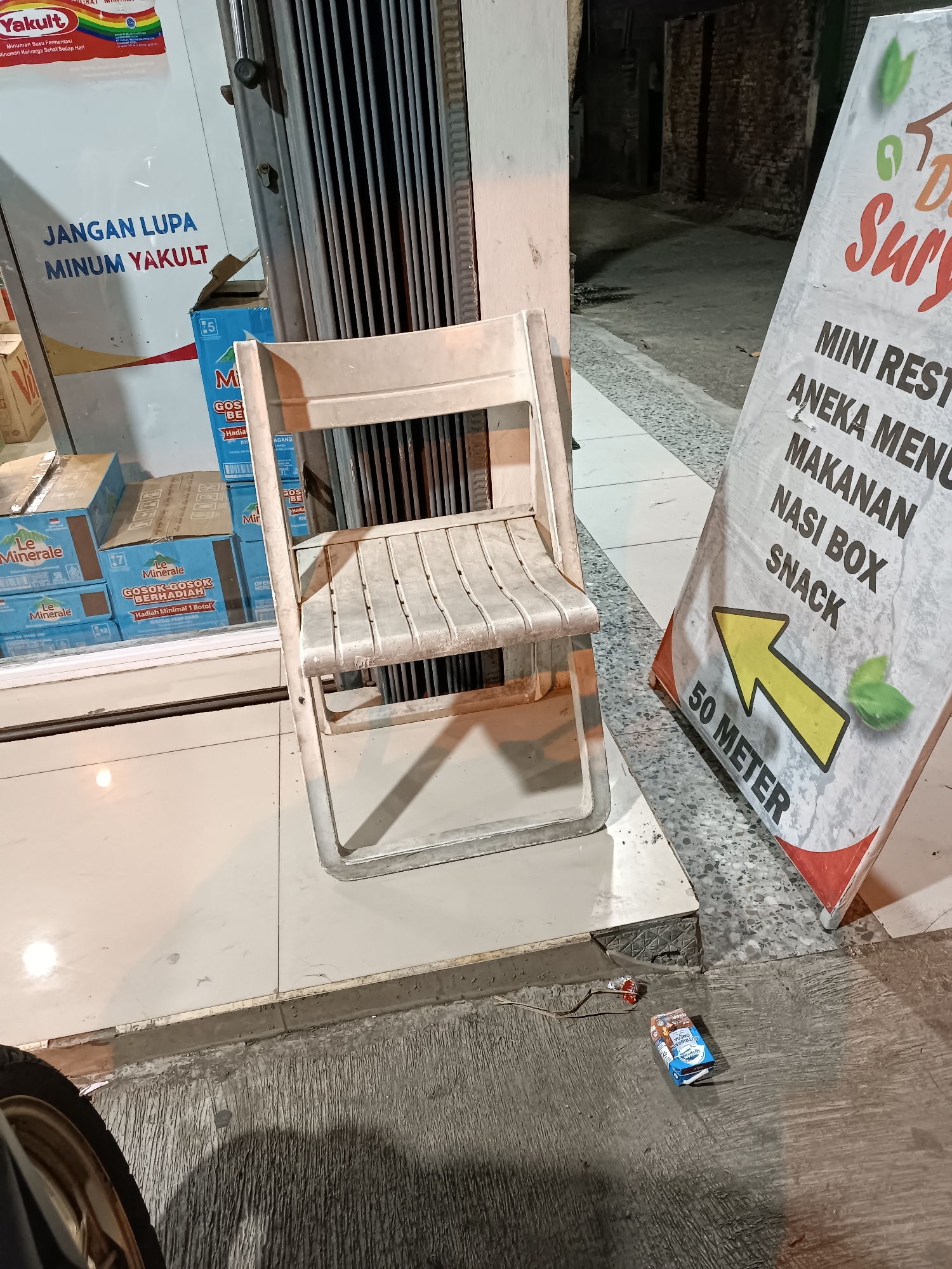 A white folding chair is placed on a tiled floor in front of a closed metal grate store entrance. To the left of the chair, a stack of bottled water boxes is visible, some with labels that read "Le Minerale" and "GOSOK-GOSOK BERHADIAH". A sign with text in Indonesian, including "MINI REST", "ANEKA MENU", "MAKANAN", "NASI BOX", and "SNACK", is leaning against the wall to the right of the chair. The sign also features a yellow arrow and the text "50 METER". On the ground in front of the chair and sign, a small carton of chocolate milk and a piece of candy are discarded. The environment appears to be an outdoor commercial area in Jetis, Indonesia, possibly a street or sidewalk. The lighting suggests it is nighttime.