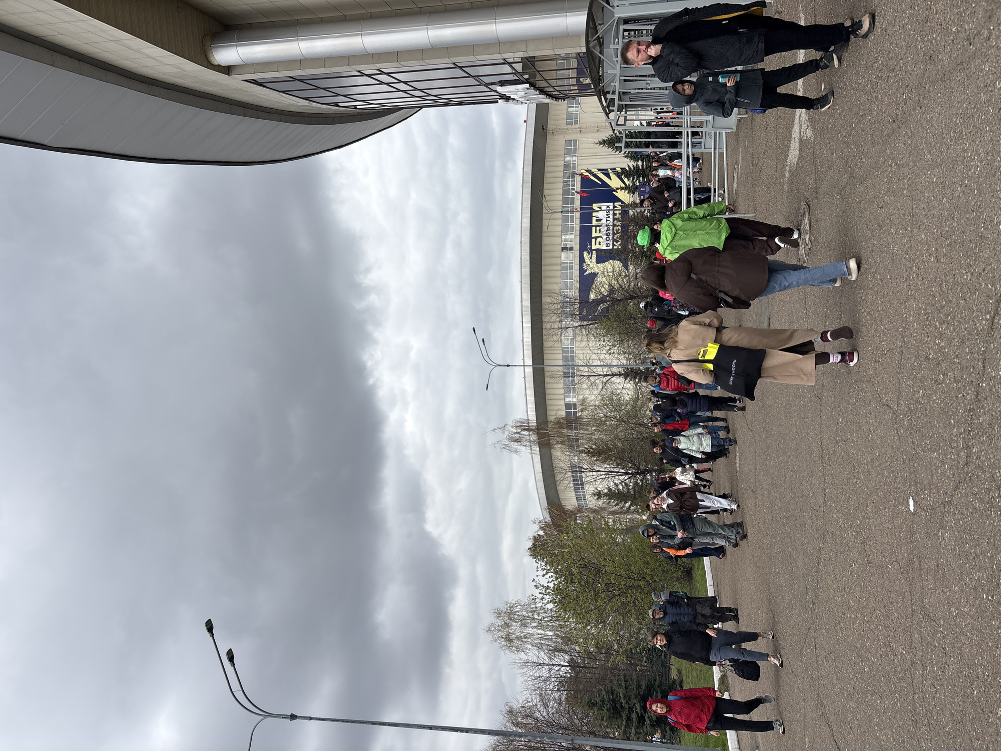 The image shows a line of people outside the TatNeft Arena in Kazan, Russia. They are waiting on a paved area in front of the building, under a cloudy sky. The arena has a modern design with a curved roof and a glass facade. A large banner is visible on the arena's facade with Russian writing that reads "Взятие Казани" (Taking of Kazan). Two streetlights stand in the foreground. People in the line are wearing various types of clothing, including coats, jackets, and hats. Some people are moving, some standing still. Some are looking at the camera, some not. Some are in groups.