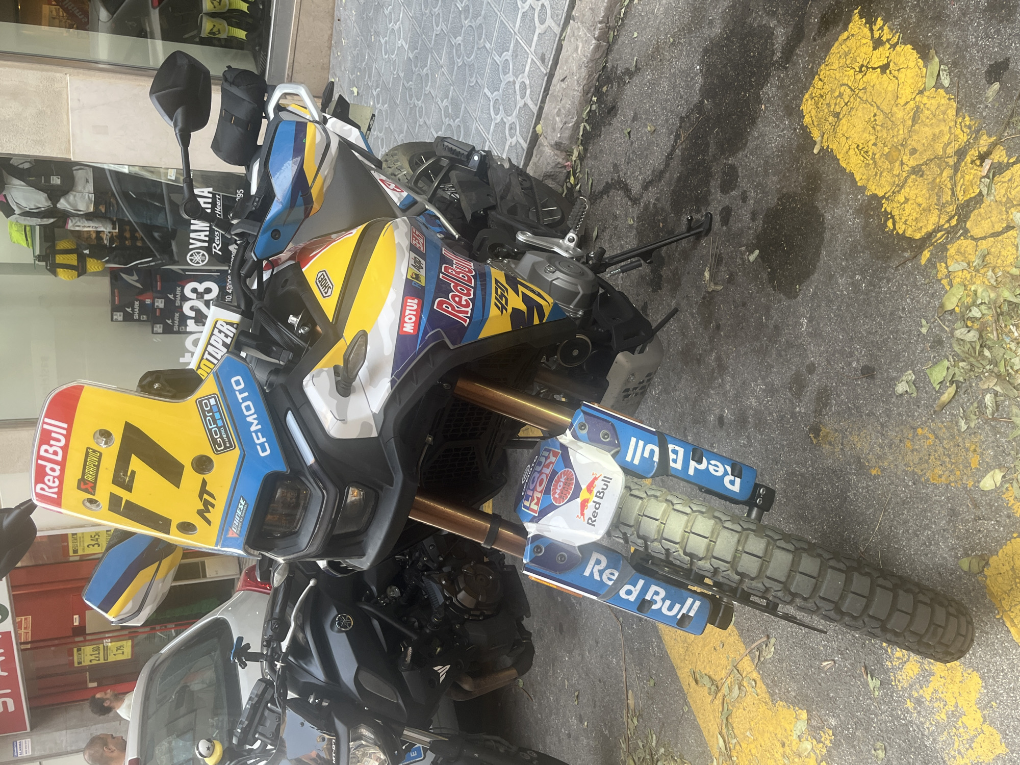 In Tarragona, Spain, on a paved street, a KTM 450 Rally motorcycle is parked. The motorcycle is adorned with racing decals, including "Red Bull" and "CFMOTO," and features a prominent yellow windscreen with the number "17." To the left of the KTM, a black Yamaha MT-07 motorcycle is partially visible. In the background, a store with display windows shows motorcycles, helmets, and other accessories. Two men are visible near a silver car on the left side of the image. The street is marked with yellow lines and scattered with fallen leaves.