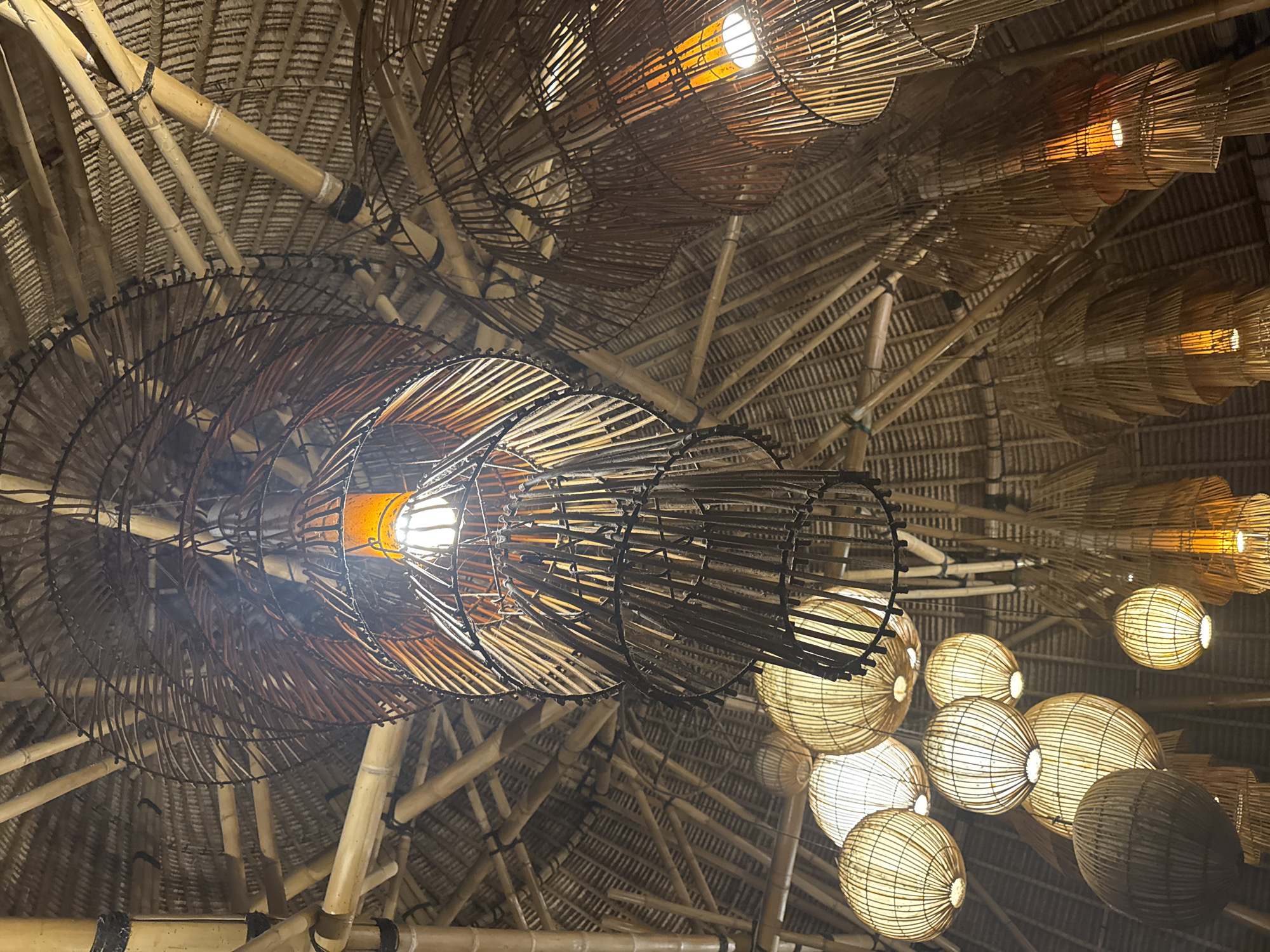 An upward-looking view captures the intricately designed ceiling of an indoor space, characteristic of tropical architecture, consistent with the Kuta Selatan, Indonesia, context. The ceiling structure is primarily composed of numerous bamboo poles arranged in a radial pattern, supporting a textured thatched roof visible between the supports.
The space is illuminated by a variety of decorative light fixtures, all contributing to a warm and inviting atmosphere. Prominently featured in the central and upper-left sections are large, elongated lanterns crafted from dark, thin strips of woven material, likely rattan or bamboo, formed into a spiral or conical shape. These fixtures contain bright light bulbs that cast a distinct, warm glow.
To the right, a cluster of spherical lanterns, made from lighter-colored woven bamboo or rattan, hang and illuminate the area with a softer, warm light. Other fixtures in the upper right resemble tiered or stacked cylindrical forms, also made of natural woven materials and emitting a similar warm light. The presence of these artificial lights suggests the image was taken at night or in an interior where such illumination is consistently utilized. The combination of natural materials like bamboo and thatch with the warm lighting creates a rustic, cozy, and exotic aesthetic. There is no visible text in the image.