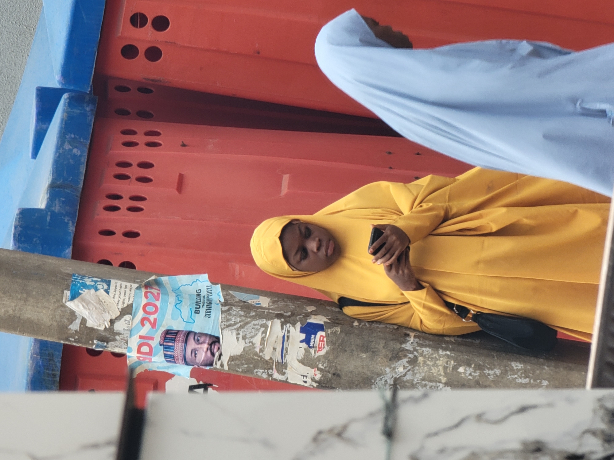In Maiburiji, Nigeria, the photograph depicts a woman in a mustard-yellow hijab standing near a concrete pillar. She is looking at a black smartphone in her hands. A second person, partially visible, wears a light blue head covering. The pillar is plastered with a ripped election poster of a candidate named NDI 2023 with the slogan "Building through serving people," along with other tattered ads and scraps of paper. Behind the pillar, there's a barrier composed of orange and blue sections with visible holes, potentially indicating construction or road management. The foreground contains a blurry white, possibly tiled, surface.