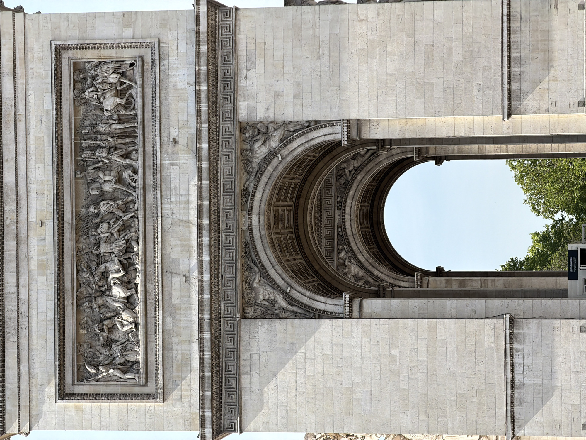 The image shows a portion of the Arc de Triomphe in Paris, France, from a low angle. The facade is constructed from large, light-colored stone blocks. The left side of the frame features a tall rectangular relief sculpture depicting a battle scene. Adjacent to the sculpture, there is a vertical strip decorated with a Greek key pattern. The archway of the monument is visible on the right, with intricate carvings above the arch. Trees and clear sky are visible through the archway. A small street sign can be seen near the trees, just outside of the monument.