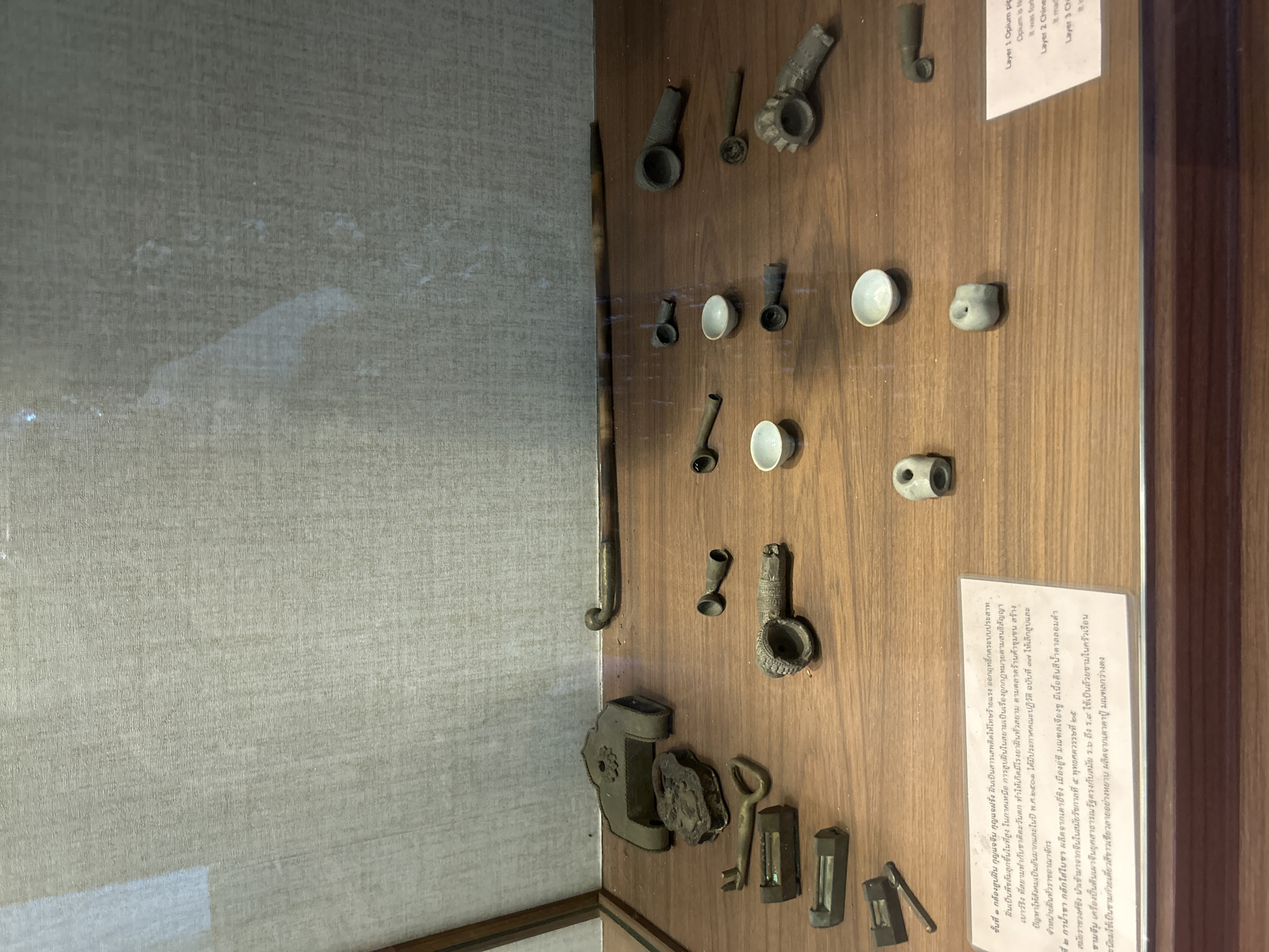 The image displays a museum exhibit within a glass case, featuring a collection of antique objects on a wooden surface. In the upper left quadrant, there are several metal objects, including what appears to be a padlock with its key and a decorative lock plate. To the right of these, a long, slender object, possibly a pipe or cane, is positioned horizontally.
The main display area is a collection of smaller items, predominantly metal and ceramic, arranged in rows. Several pipe-like objects, some with intricate detailing, are visible, alongside small ceramic bowls or cups. The items are laid out in a way that suggests they are artifacts from different layers or contexts, as indicated by a small label in the lower right corner that reads "Layer 1 Opium pipe", "Layer 2 Chinese", and "Layer 3 Chi".
A partially visible text in Thai is present in the lower left portion of the image, likely providing information about the artifacts. The background is a textured, light-colored wall, possibly fabric or wallpaper. The overall lighting is even, suggesting an indoor display setting. There are no people or active interactions depicted in the scene. The environment is a controlled museum display. The location is not explicitly stated in the visible text or visual cues within the image, other than it being an exhibit.