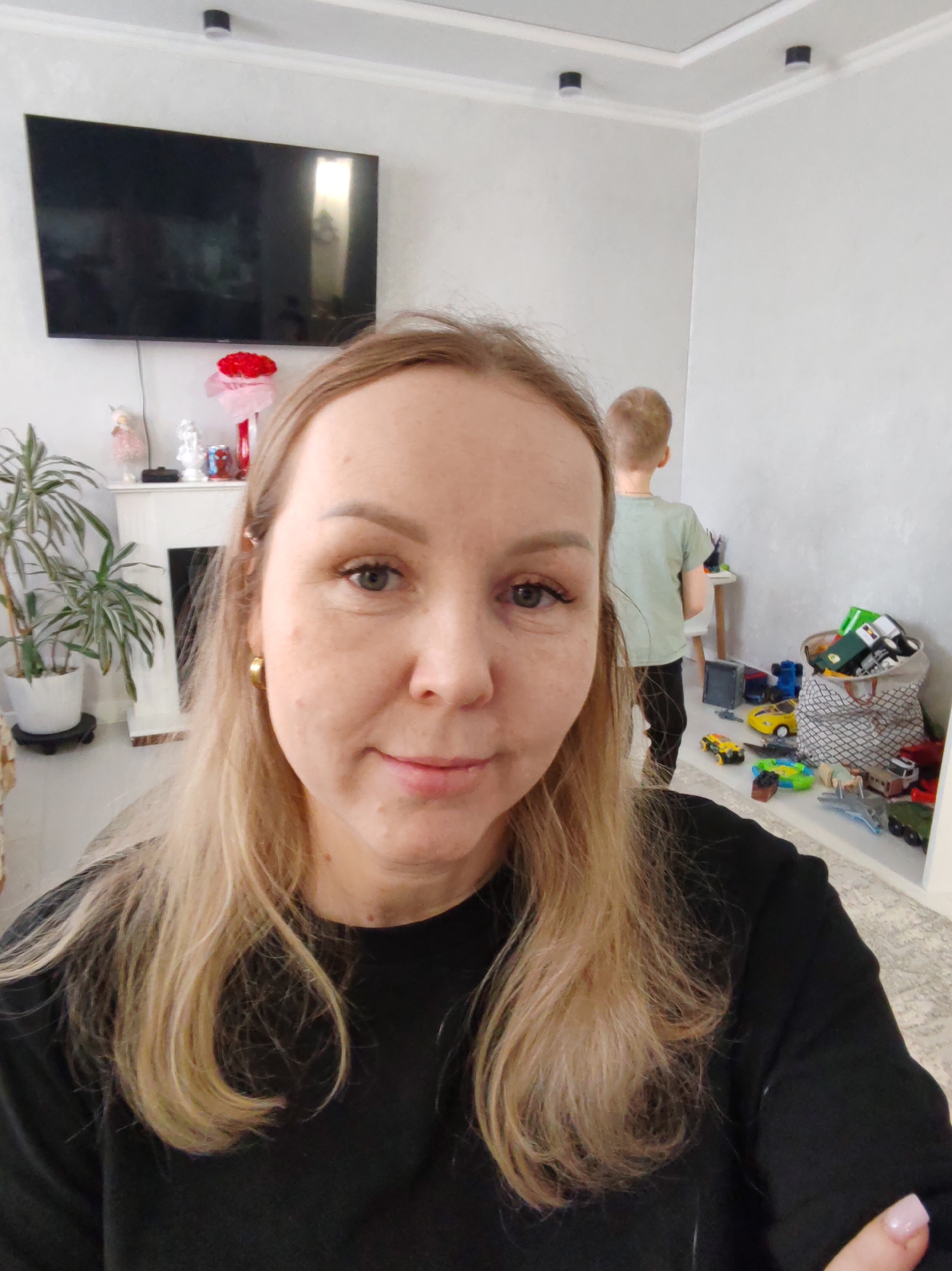 A woman with light brown hair and a black long-sleeved shirt is taking a selfie in a room. Behind her, a young boy with short blonde hair is standing near a white table. Next to the boy is a collection of toys on the floor and in a patterned basket. To the left of the woman, there is a plant in a white pot and a white mantelpiece with decorations, including a bouquet of red flowers. A large black television is mounted on the wall above the mantelpiece. The walls are painted a light gray color. The ceiling is white with recessed lighting. Based on the context, this location is likely within a residential setting. The mention of Chaikovsky, Russia, is likely relevant to the location.