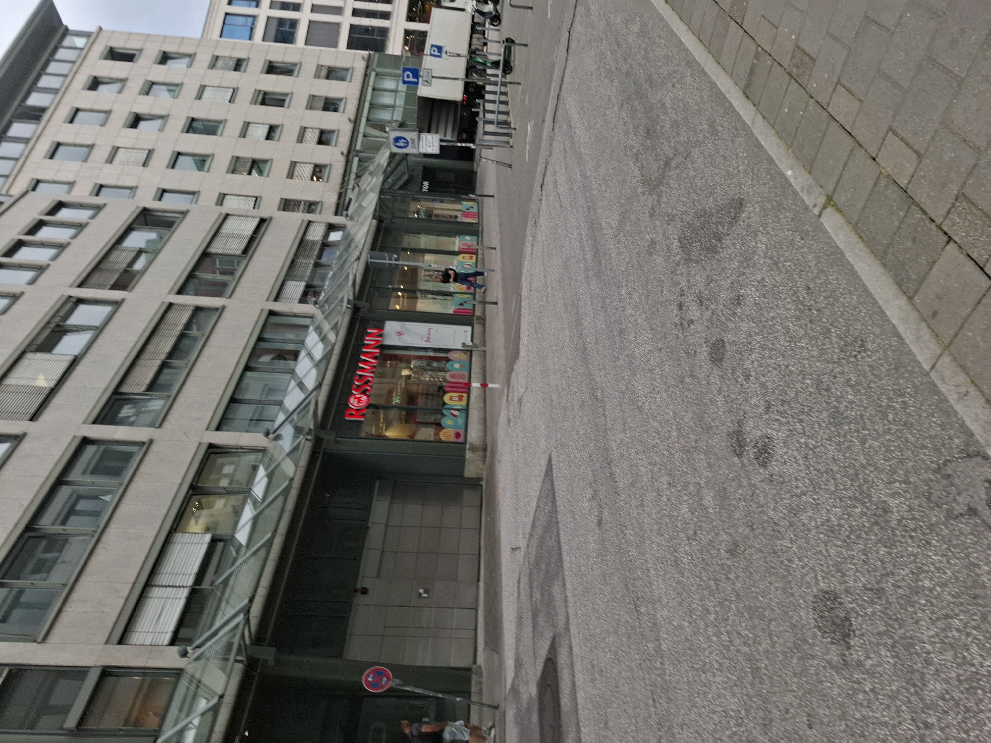 The image depicts a street scene in Hamburg, Germany, in front of a Rossmann drugstore. In the foreground, a paved road with some dark stains is visible, transitioning to a sidewalk made of grey rectangular pavers on the right. A person in a dark t-shirt and shorts is walking on the sidewalk to the left of the frame. Adjacent to the sidewalk is a street with parked bicycles and a white truck. A sign with a "P" indicates parking. Further back, the facade of a modern building is visible, featuring multiple floors with numerous windows. The ground floor hosts the Rossmann store, recognizable by its red illuminated signage. A glass canopy extends over the entrance. A man in a dark t-shirt and blue jeans is walking past the store entrance. A red and white striped pole stands to the right of the store. In the background, other buildings and a cloudy sky are visible.