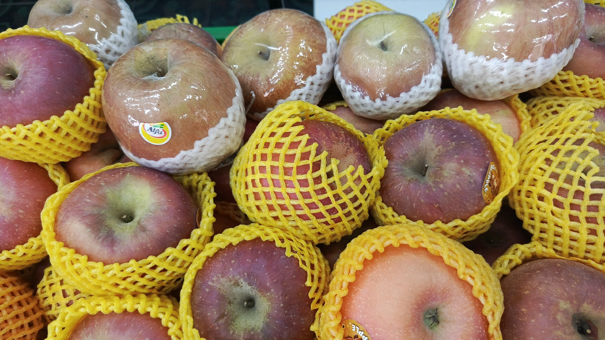 A close-up, high-angle shot shows a pile of red and pink apples at a market stall in Mangunsuman, Indonesia. The apples are individually wrapped in protective plastic and some are nestled in yellow foam netting. The lighting is bright, suggesting an indoor or covered outdoor setting. One apple in the center has a sticker with a green and red logo that reads "Alfa". The apples appear fresh and are arranged in a visually appealing manner, likely for sale.