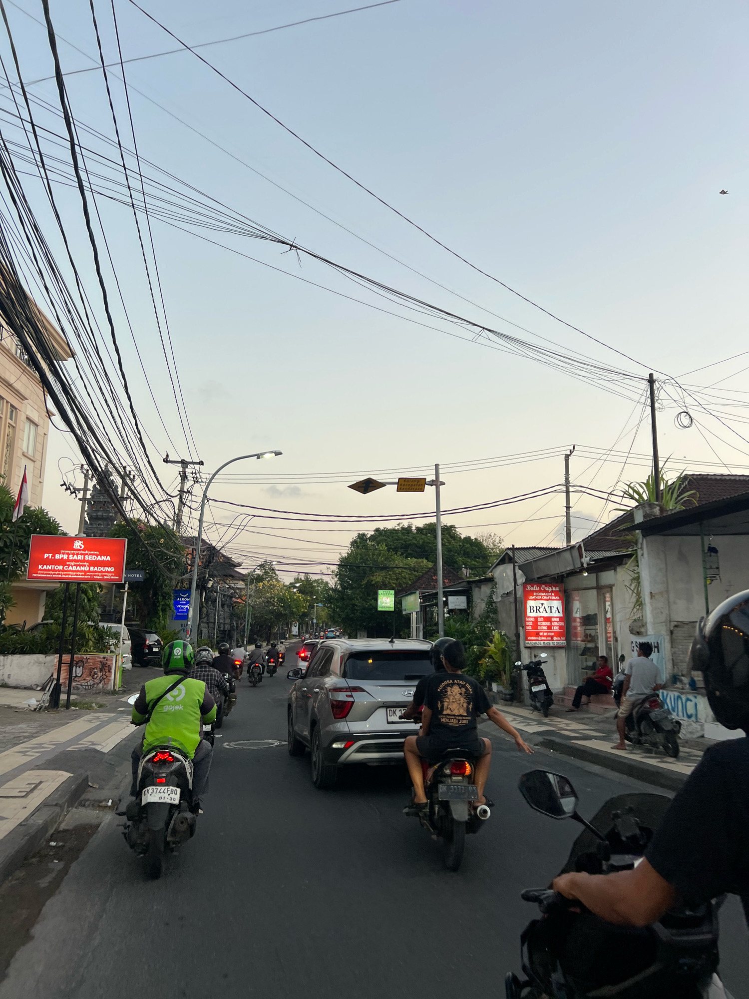 The image depicts a street scene in Kuta, Indonesia, under a hazy sky that suggests either late afternoon or early morning. The foreground is dominated by a busy road with several motorbikes and a car. A person wearing a bright green Gojek jacket is riding a motorbike in the left lane. Further down the road, other motorbikes are visible, some with passengers. A silver SUV is in the right lane.
The environment is characterized by buildings on both sides of the street. On the left, a multi-story building with a sign that reads "PT. BPR SARI SEDANA KANTOR CABANG BADUNG" is prominent. The buildings on the right appear to be shops, with one displaying a sign for "Baliz Origina BRATA" which appears to be a shoemaker. The street is lined with trees and power lines, which are numerous and strung across the sky, adding to the urban feel. There are also streetlights and traffic signals. The overall impression is of a typical, bustling street in a Southeast Asian city.