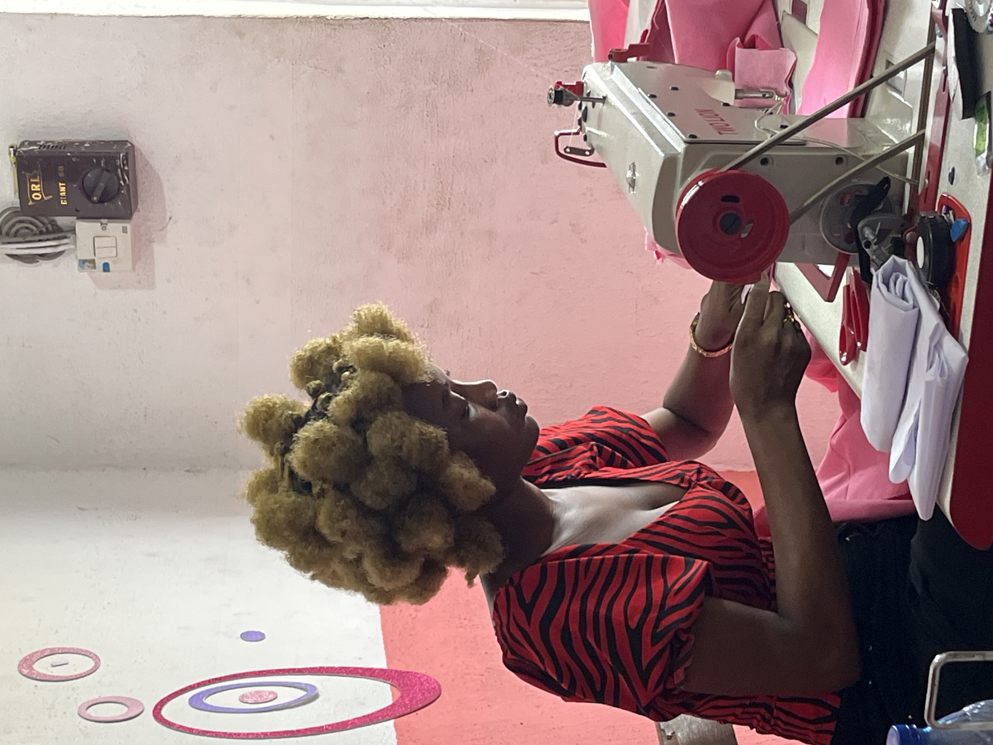 In an indoor setting in Badagry, Nigeria, a woman with afro-style hair stands next to a sewing machine, operating or adjusting it. The woman wears a red and black striped shirt and dark pants. The sewing machine is light-colored with red components and is surrounded by pink fabric. A light switch and electrical box are visible on the wall. The floor has painted decorative circles.