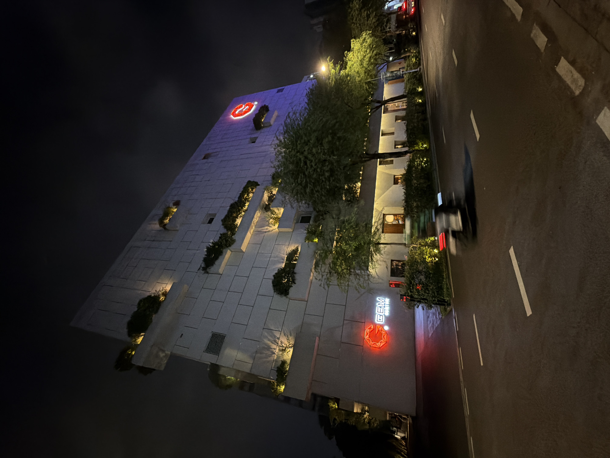 At night, an exterior shot shows a building in Thủ Đức, Vietnam. The building has a white facade with staggered tiers of greenery. Red neon signs are visible on the front and top. A tree lines the building. To the right is a street with white lane markers. A vehicle, blurry due to motion, is present. The sky is dark.