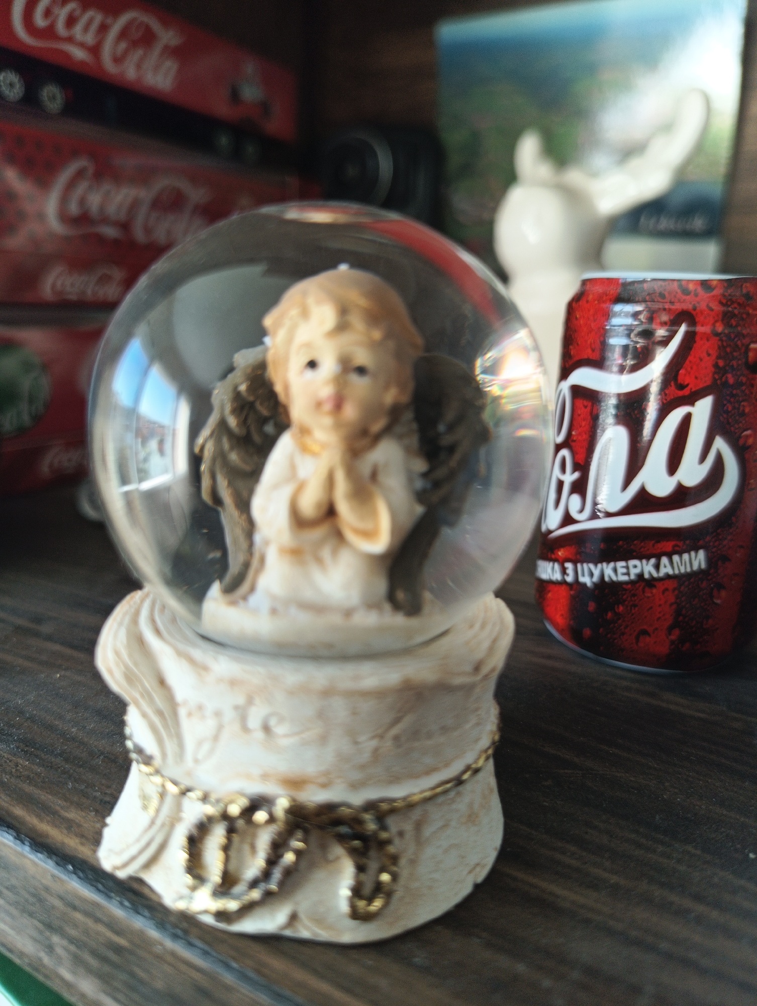 A close-up, eye-level shot shows a decorative snow globe with an angel figurine inside, placed on a dark wooden surface. The snow globe has a clear glass dome and a cream-colored base adorned with a gold chain. Behind the snow globe, a red Coca-Cola can with condensation is visible. In the background, stacked Coca-Cola boxes are blurred, and a white statue with outstretched arms is partially visible against a colorful, out-of-focus backdrop. The scene is likely an indoor display, possibly in a home or shop, in Karnobat, Bulgaria.