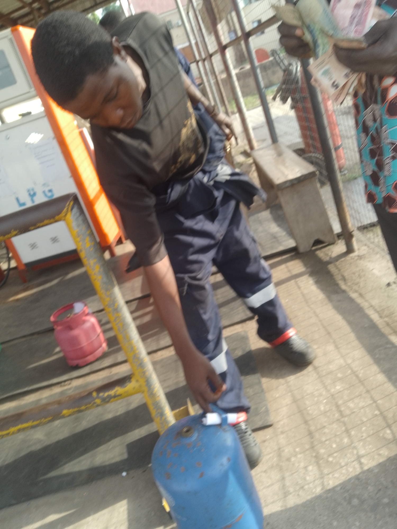 A man in a dark t-shirt and navy work pants is tightening a valve on a blue gas tank in an outdoor setting. Next to him is a smaller red gas tank. The background includes a bench, a yellow and grey metal bar, and another person holding money. There is also an 'LPG' sign visible. The location is in the city of Unknown, Unknown.