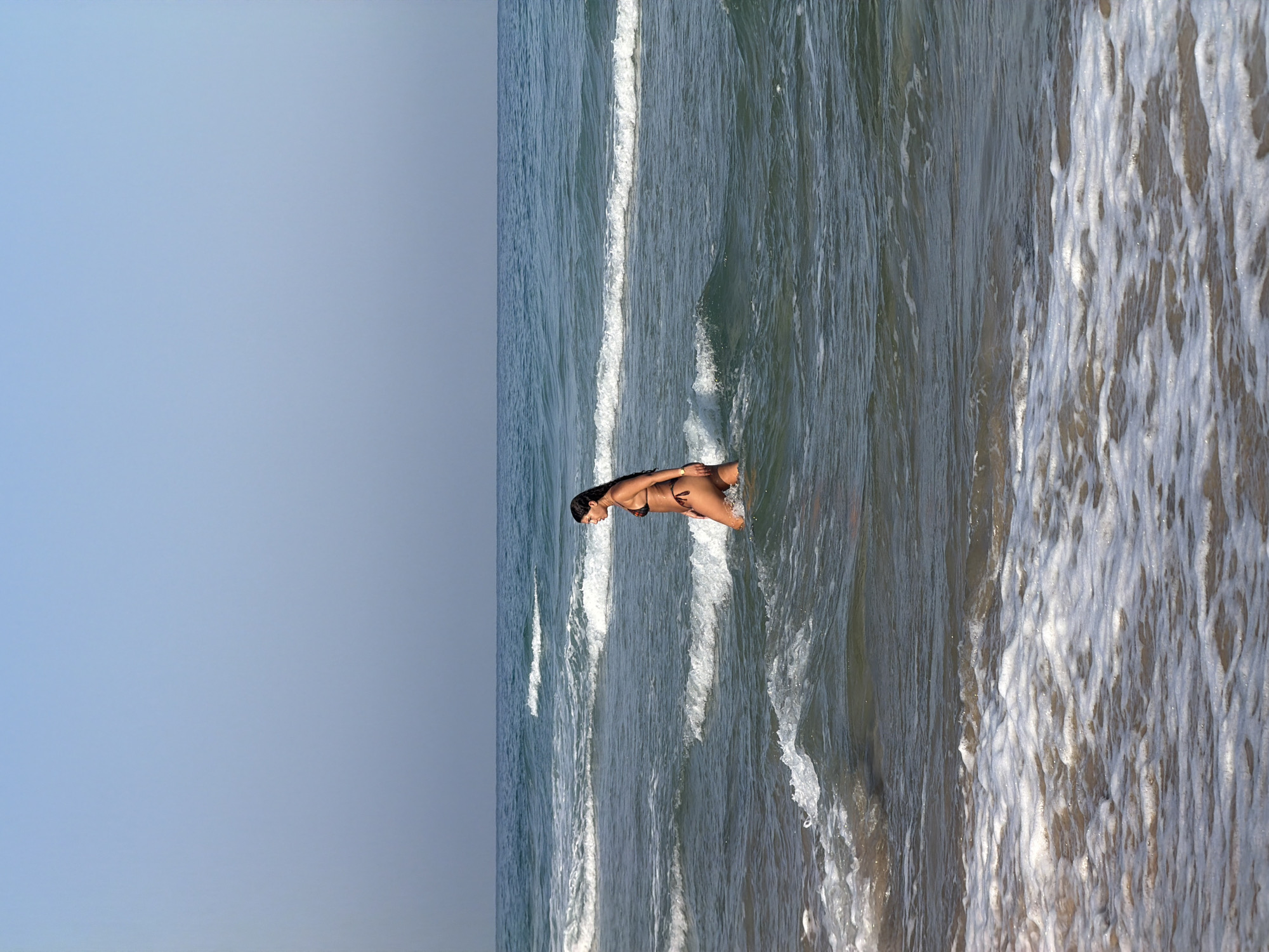 A woman is wading in the shallow, clear ocean water near the shore. The woman is wearing a dark-colored bikini and has long, dark hair. She is facing to her left, with waves breaking around her legs. The water is a light blue-green, and the sky above is a pale blue with no clouds. The background shows a hazy horizon where the sea meets the sky. The image appears to be taken during the day with natural light. The scene is likely on a beach in Bat Yam, Israel, given the context.