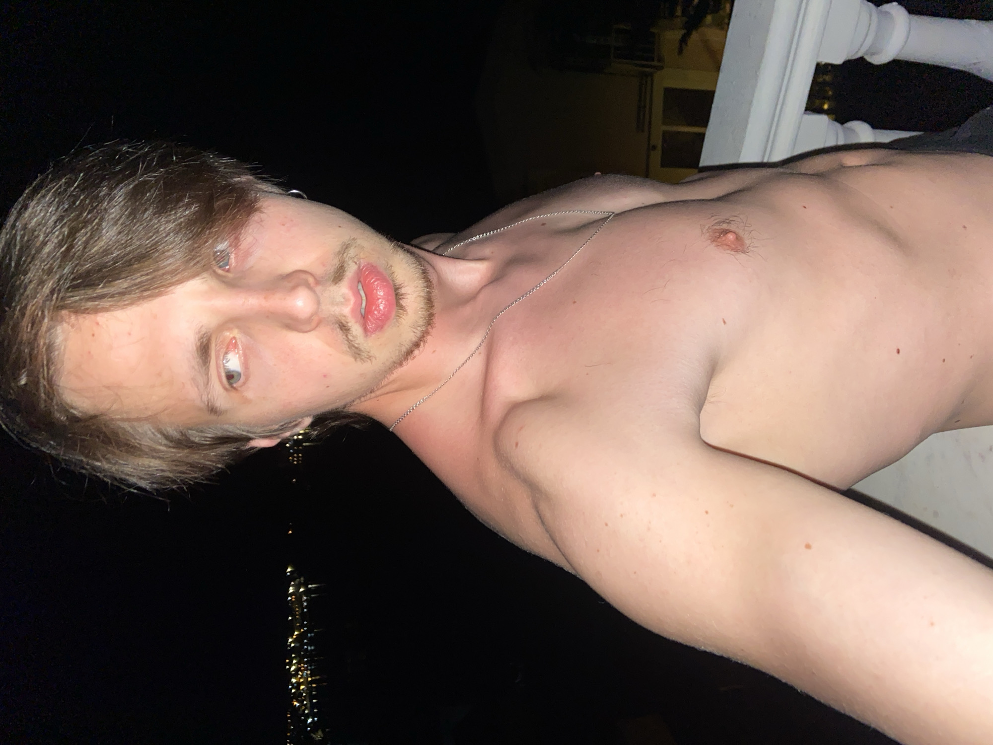 A shirtless young man with brown hair and light eyes poses for a selfie. He wears a thin silver chain around his neck. The background shows a dark sky, with the lights of Šušanj, Montenegro visible in the distance. A white railing is partially visible in the upper right corner.