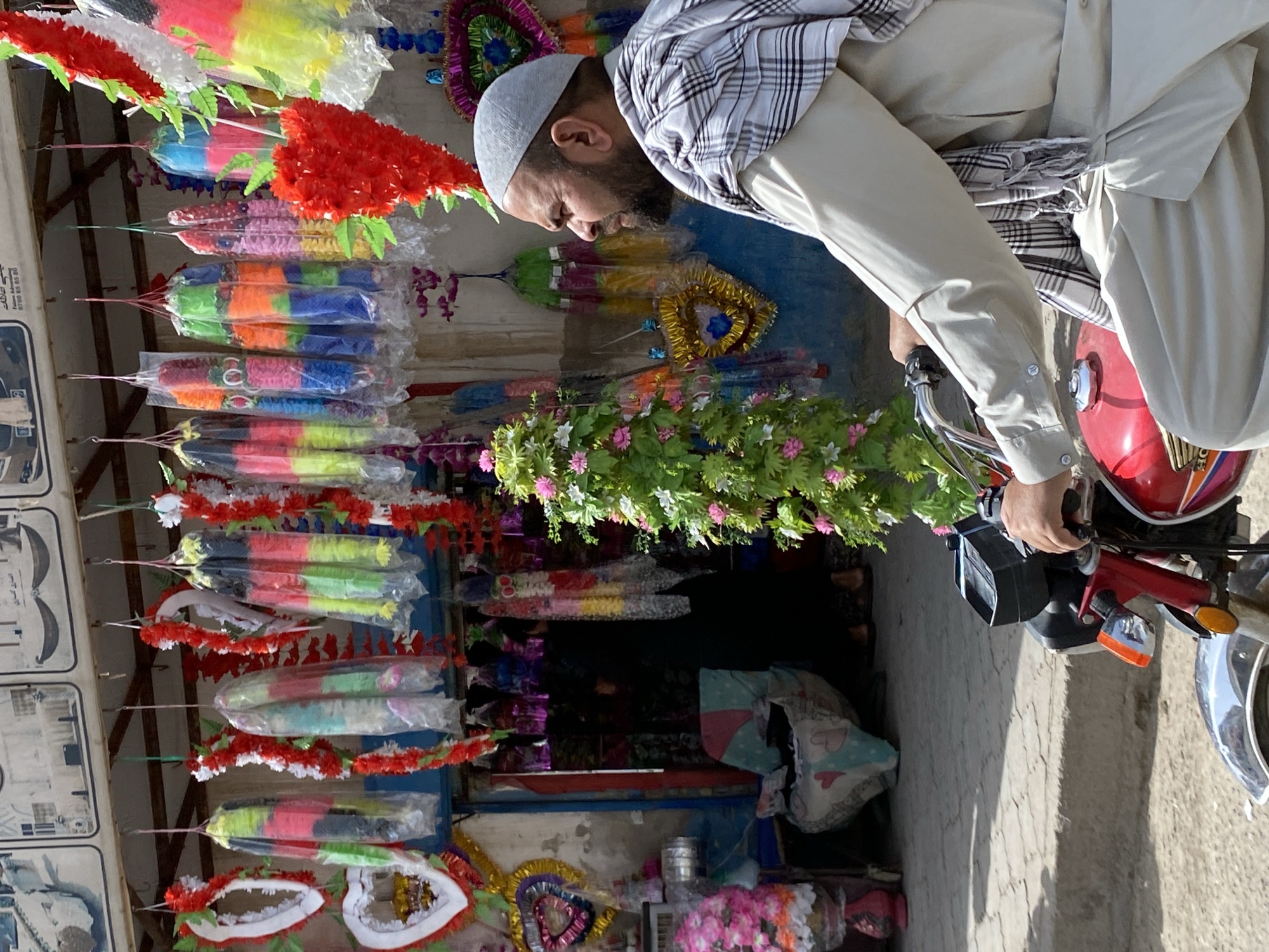 The image depicts a street scene in Kandahar, Afghanistan, outside a shop adorned with various colorful festive decorations. In the foreground, a man wearing a light-colored tunic and a checkered scarf is seated on a red motorcycle. He has a beard and is looking to the right of the frame. The motorcycle has a black box attached to the handlebars. Behind the man and the motorcycle, the shop displays an array of hanging decorations, including colorful garlands and heart-shaped ornaments, made of plastic and fabric. Some of these decorations are wrapped in plastic. A green, leafy artificial plant with pink flowers stands to the left of the man. Partially visible inside the shop is a person in dark clothing. The ground in front of the shop is paved with rough stones. The overall impression is one of a vibrant commercial area with celebratory merchandise on display.