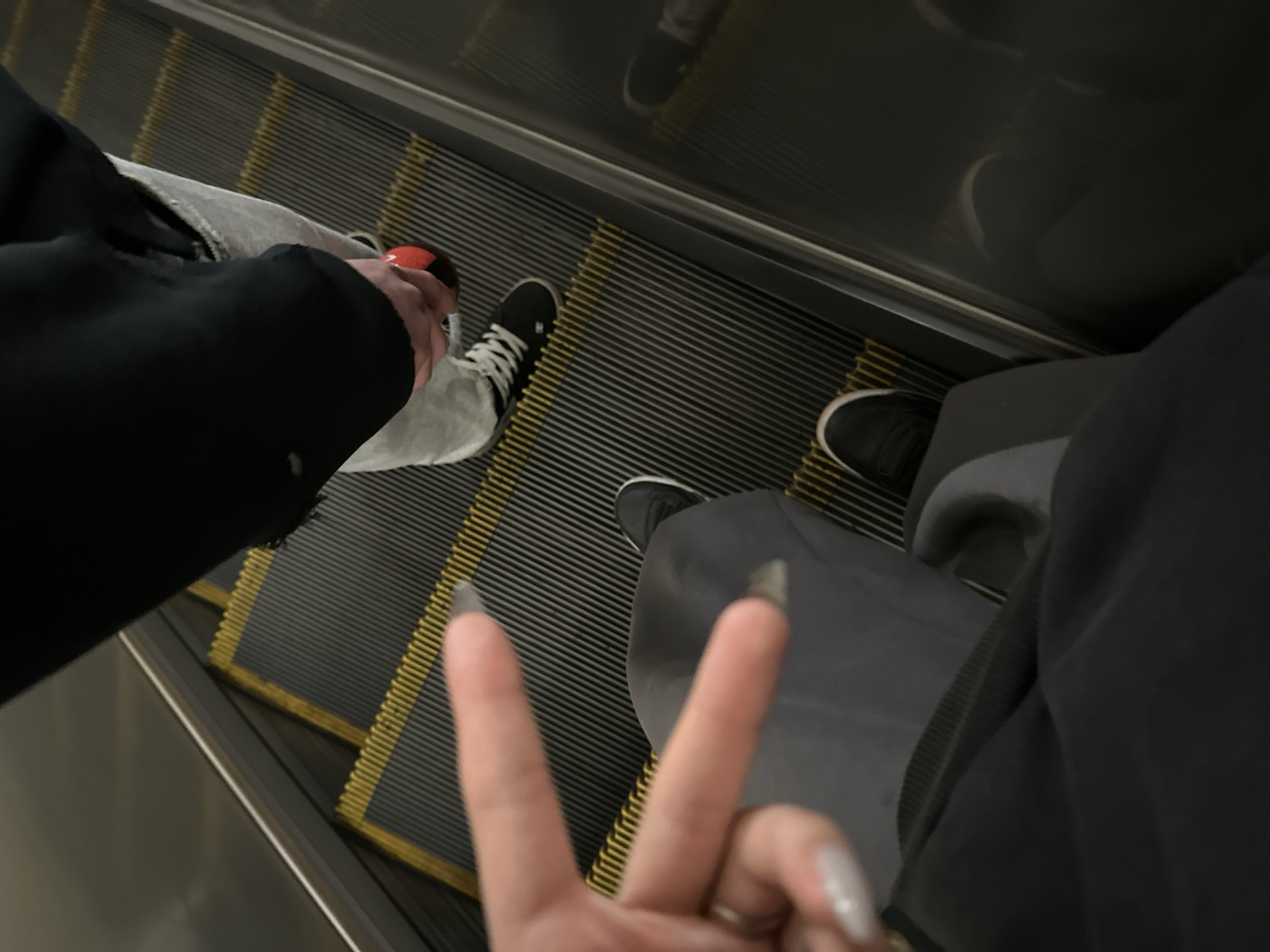 A low-angle shot shows an escalator ride shared by two individuals. The person on the left wears grey denim pants, a black hoodie, and black-and-white sneakers with white laces and a red accent on the heel. The person on the right is wearing grey pants and dark colored sneakers. The person on the right also presents a "peace" sign with their fingers, while traveling on the escalator. The background indicates a commercial or public space. The location in the city of Moscow, Russia cannot be determined from the image.