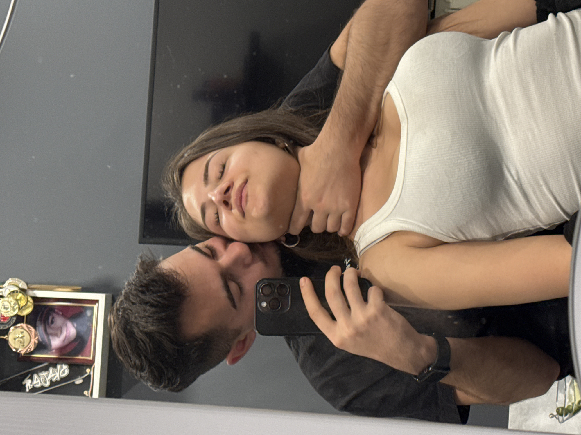 This mirror selfie, taken in Sofia, Bulgaria, features a young man and a young woman posing intimately. The image is oriented sideways, with the subjects appearing to be lying down, though it is likely a mirror reflection captured from a different angle.
The man, with short dark hair, is positioned behind the woman, holding a black smartphone with a triple camera setup in his right hand to capture their reflection. He wears a dark t-shirt and a dark watch on his left wrist. His left arm is around the woman's neck, with his hand gently positioned around her throat, a pose that appears playful or affectionate rather than aggressive, given their relaxed expressions. He leans his head towards the woman, his eyes closed.
The woman has long, dark brown hair and wears a white ribbed tank top. Her eyes are also closed, and she has a serene or peaceful expression, leaning into the man's embrace.
In the background, visible in the mirror's reflection to the left, is a shelf holding a framed picture of a person, possibly in a uniform or costume, and several medals or decorative items. The word "RAFAYO" is visible on the frame. A large dark screen, possibly a TV, is also reflected in the upper part of the mirror. The room appears to have dark gray walls. The overall mood is intimate and affectionate.