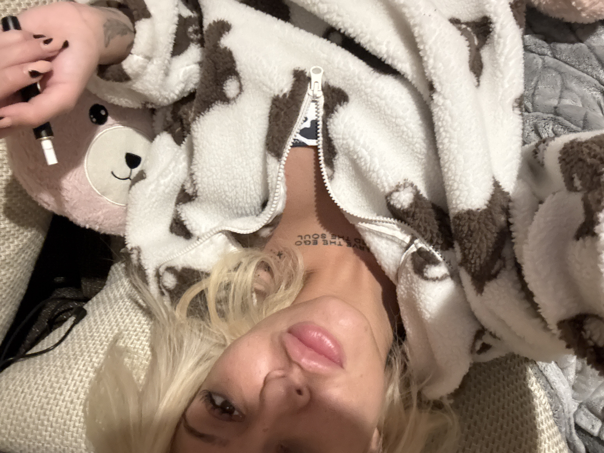 A woman with blonde hair is lying down, looking directly at the camera. She is wearing a fluffy white and brown spotted (possibly cow print) zipped hoodie or onesie. Her hair is spread around her head on a light-colored, knitted blanket. Her lips are slightly parted, and she has dark eyeliner.
To her left, a pink teddy bear plushie is visible, with a small portion of its face showing. The woman's hand, adorned with dark nail polish, holds a black and white pen-like object (possibly a vape pen) above the teddy bear. A tattoo is visible on her neck, reading "SAVE THE EGO" on the top line and "AND THE SOUL" below it, though it appears inverted from the viewer's perspective.
The background consists of various textile textures, including the knitted blanket she is lying on and a quilted, grey blanket to her right. The overall setting suggests a casual, indoor environment. The lighting is soft, indicating an indoor scene, possibly during the evening or night. The activity appears to be a relaxed selfie moment. The location context is Plovdiv, Bulgaria.