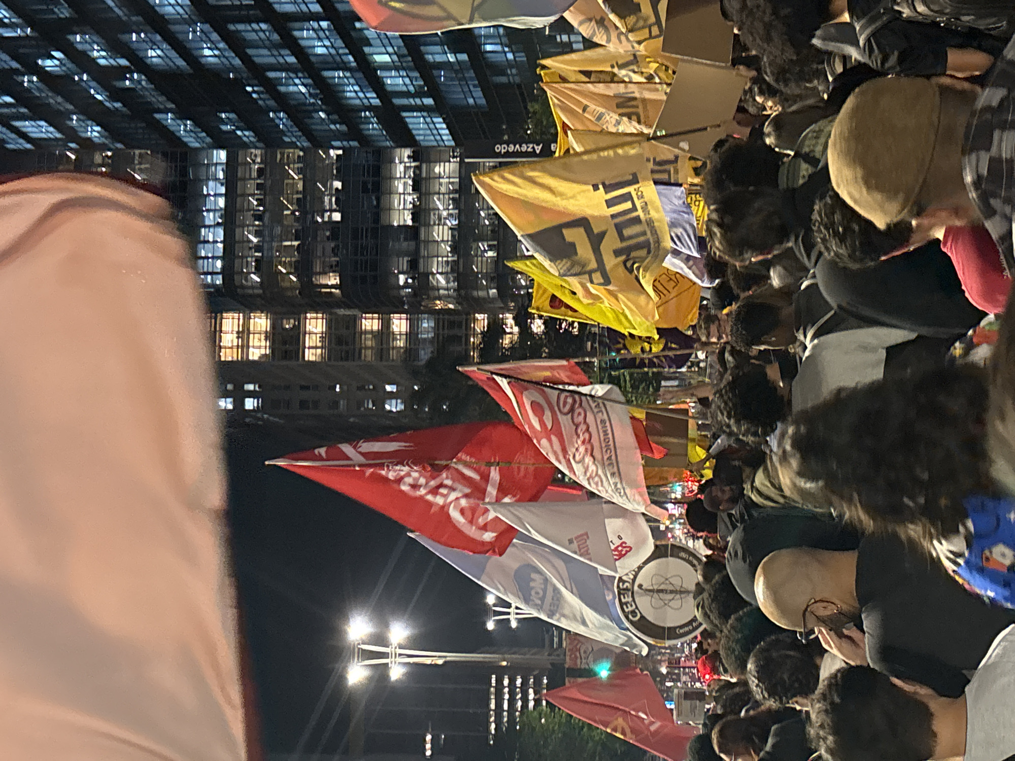 Captured at night in São Paulo, Brazil, the image depicts a large crowd of people in a public area, likely a street or plaza. A high-rise building with numerous lit windows provides an urban backdrop, its architecture reflecting the city's skyline. Visible signage includes "Azevedo." A multitude of flags and banners are being held aloft by the crowd. People are standing close together, suggesting a demonstration or gathering. The lighting is a mix of streetlights and illumination from the surrounding buildings.