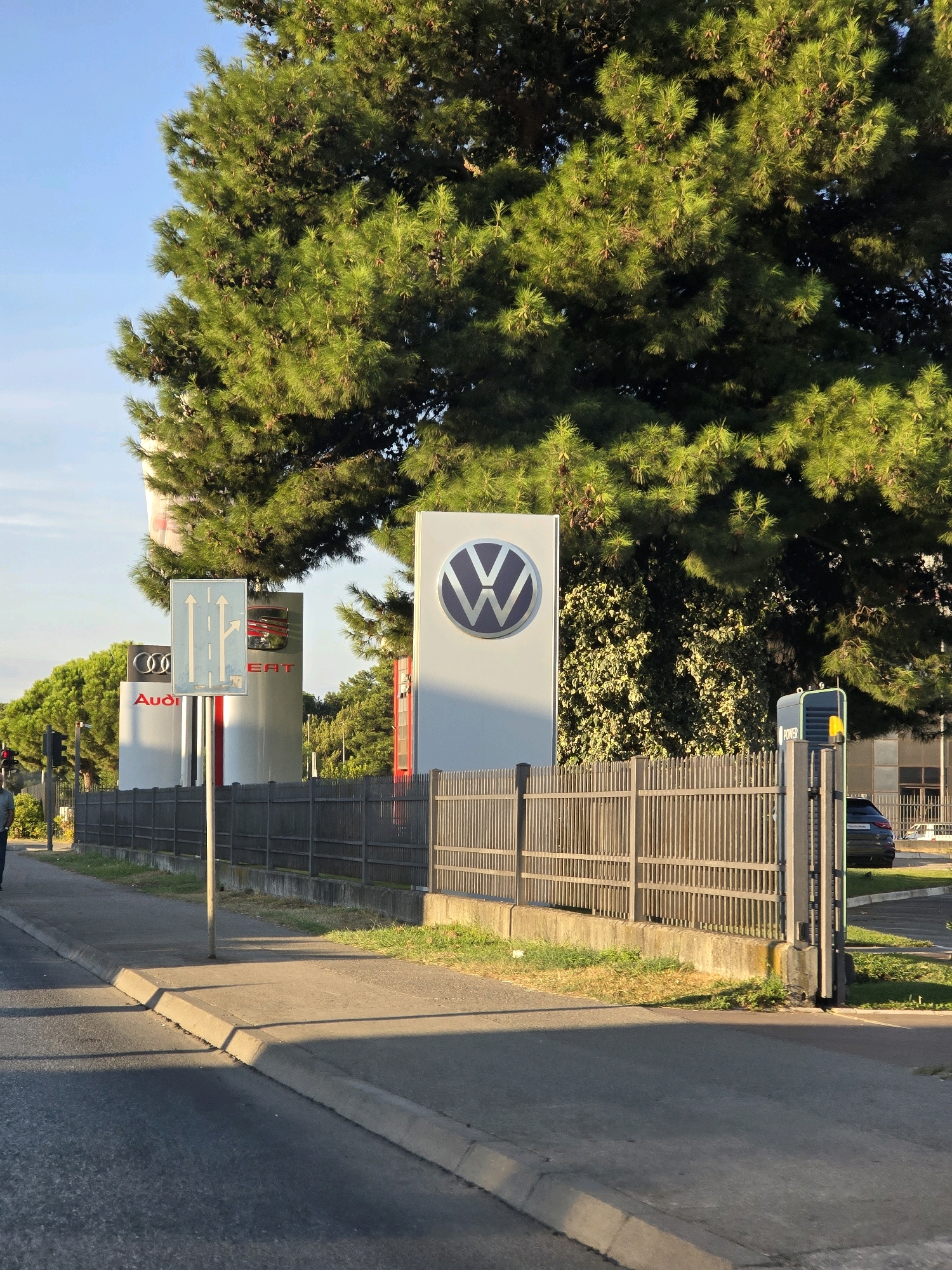 The image depicts a street scene with a car dealership entrance. A tall evergreen tree with cones dominates the upper half of the frame. In the middle ground, there are signs for car brands, including Audi and Volkswagen. A traffic sign with arrows indicating straight and right turns is positioned in front of the signs. A metal fence runs along the property line, separating the sidewalk from the dealership. A modern electric vehicle charging station is visible to the right of the Volkswagen sign. In the background, a dark-colored car is parked within the dealership lot. The ground consists of a paved road and sidewalk, with a curb separating the two. The lighting suggests it is either early morning or late afternoon, with long shadows cast across the scene. The location is identified as Podgorica, Montenegro.