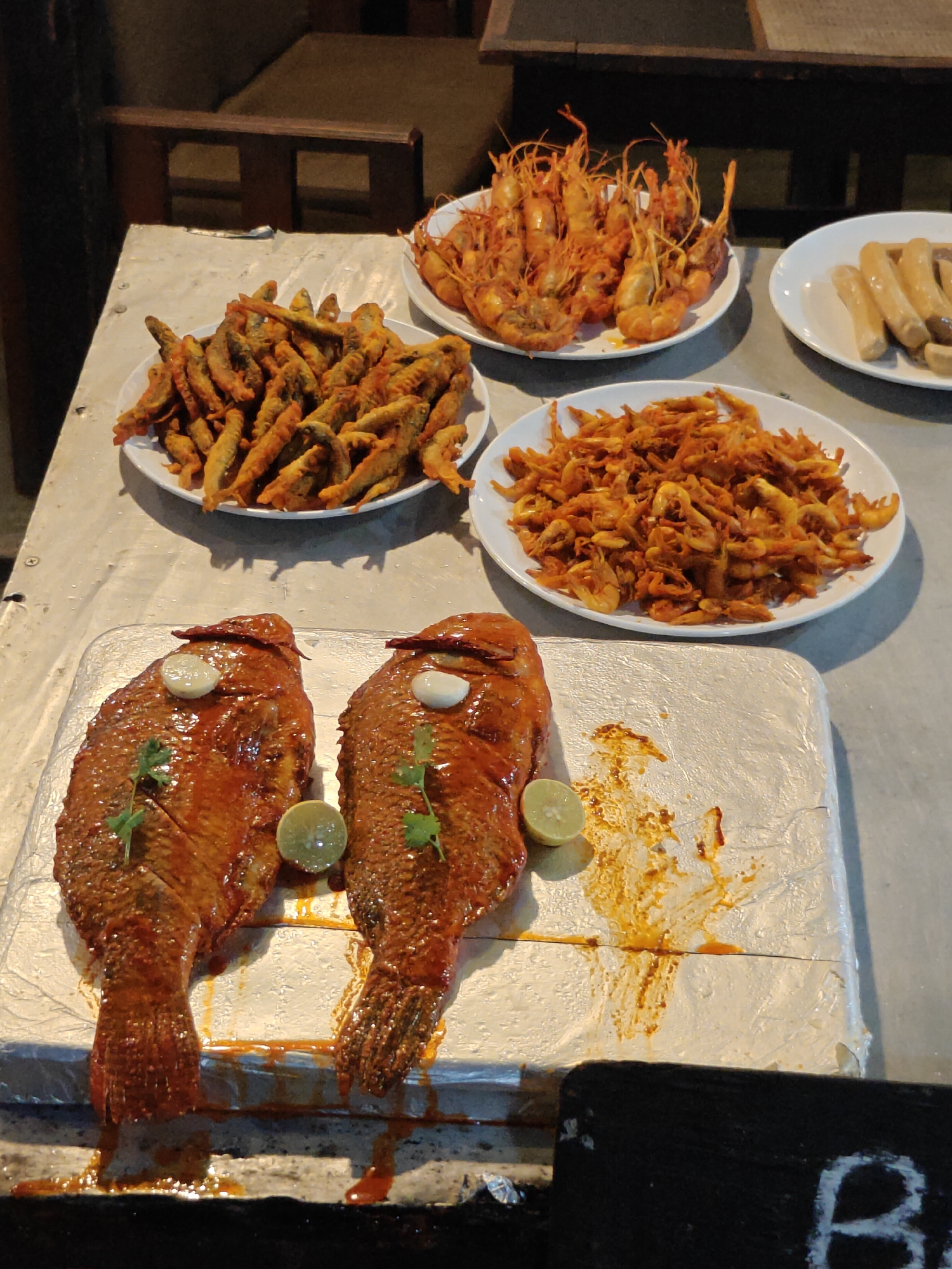 In Pokhara, Nepal, a food stall displays a variety of seafood and sausages. Two whole fish, glazed with a reddish sauce, are presented on a silver tray. Garnishes of lime wedges, cilantro sprigs, and red chilies adorn the fish. Behind the fish, three plates are filled with fried delicacies. One plate contains an assortment of small, fried fish. Another plate is piled high with fried shrimp, their shells a vibrant orange. The third plate holds several cooked sausages. The stall is set up on a textured surface, possibly a table, with wooden chairs visible in the background.