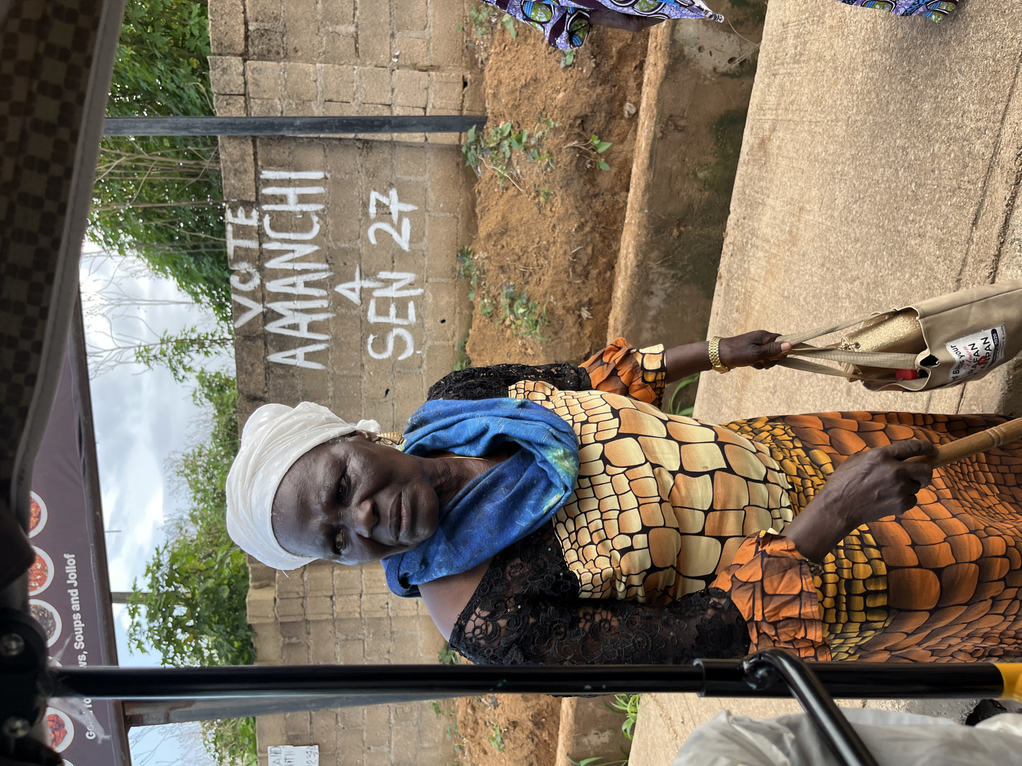 An elderly Black woman wearing a white headscarf, a blue scarf, and a colorful dress with a black lace overlay stands in front of a concrete wall with graffiti. The graffiti reads "VOTE AMANCHI 4 SEN 27". She is holding a light brown tote bag with gold accents. The woman is looking to her left, with her mouth slightly open. In the background, there are green trees and shrubs, and a dirt embankment. The scene appears to be outdoors, likely in a street or public area. The image is framed with what looks like part of a vehicle or structure on the left side. A portion of another person in a patterned garment is visible on the right edge of the frame. The overall lighting suggests it is daytime, with an overcast sky. The location is identified as Bukuru, Nigeria.