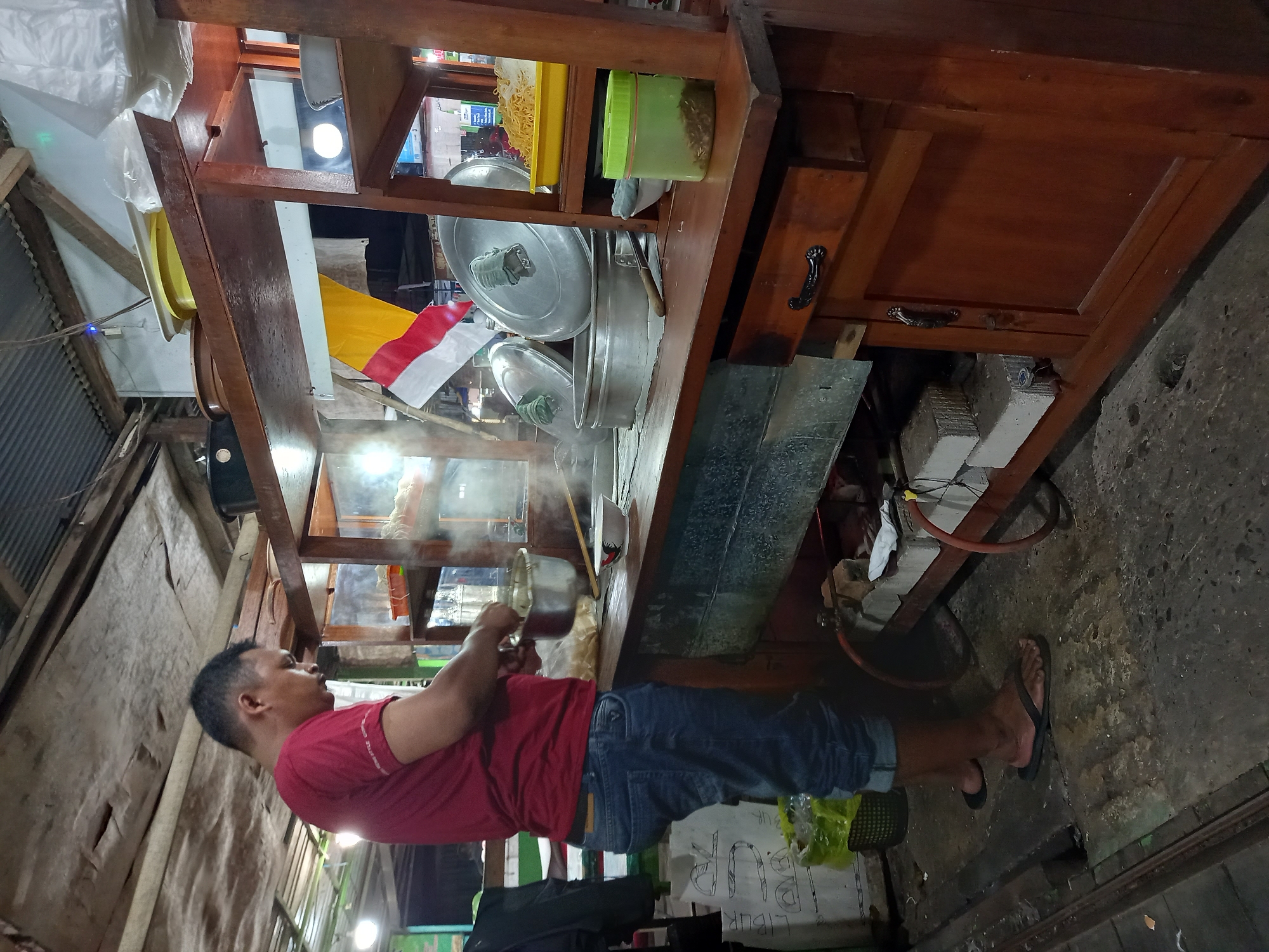 A man in a red t-shirt and blue jeans is standing at a food stall, cooking. He is holding a metal pot, and there is steam rising from the stove. The food stall is made of wood and metal, and it is stocked with various ingredients and cooking utensils. In the background, there is a flag with red, white, and yellow stripes. The scene appears to be in an outdoor market in Samarinda, Indonesia, given the tropical climate, local architecture, and the presence of street food vendors. The lighting suggests it is either early morning or late evening.
