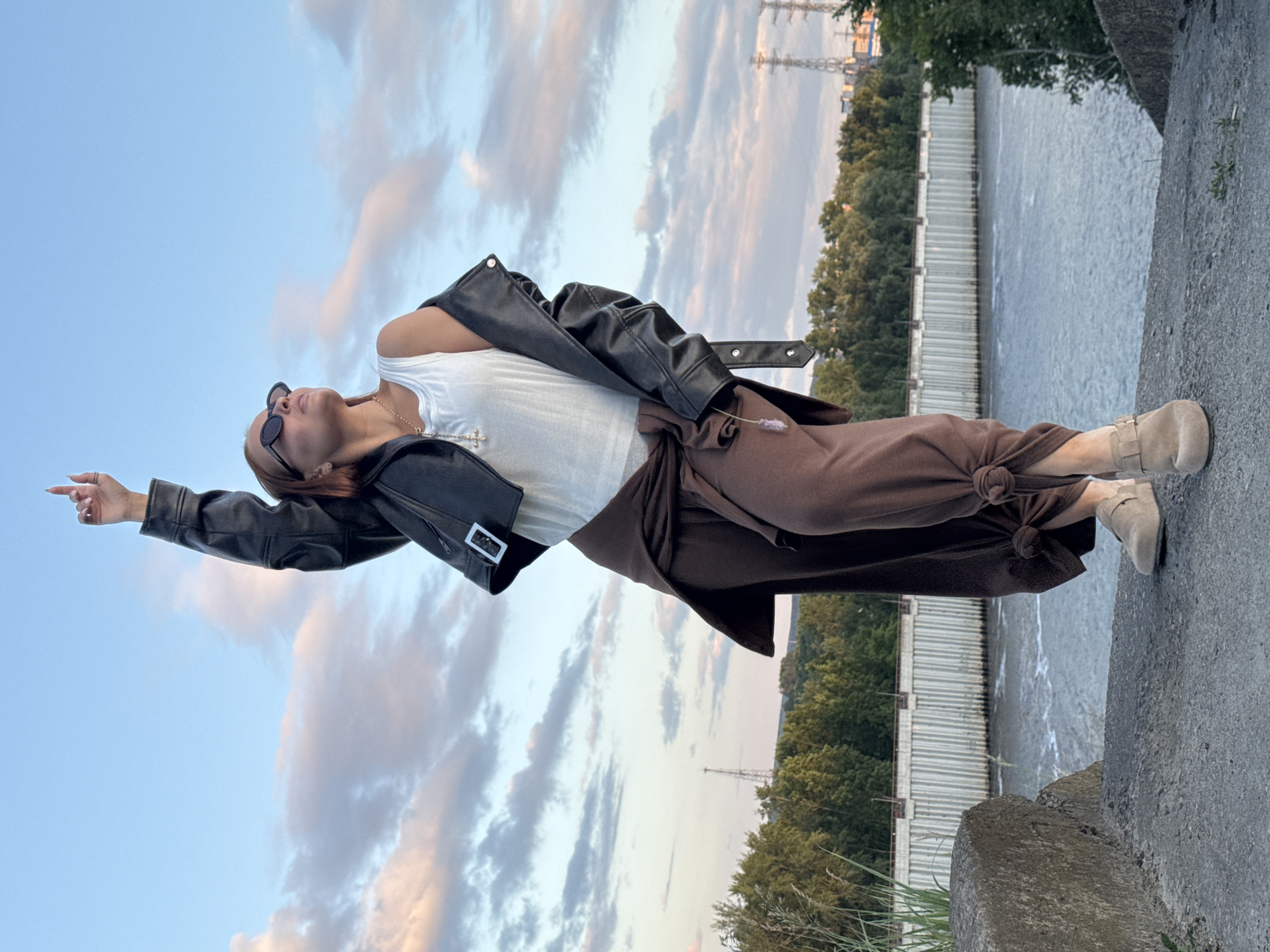 A person stands on a concrete embankment next to a river, with their right arm raised and index finger pointing upwards. They are wearing a white tank top, a black leather jacket slung over their shoulders, and loose brown pants tied at the ankles. Sunglasses are worn on their face. The background features a wide river, a concrete retaining wall with vertical metal supports, a line of trees, and a sky with scattered clouds. The scene appears to be in Chaikovsky, Russia, possibly near the Kama River dam or a similar riverside industrial area, given the presence of industrial structures and the wide expanse of water.